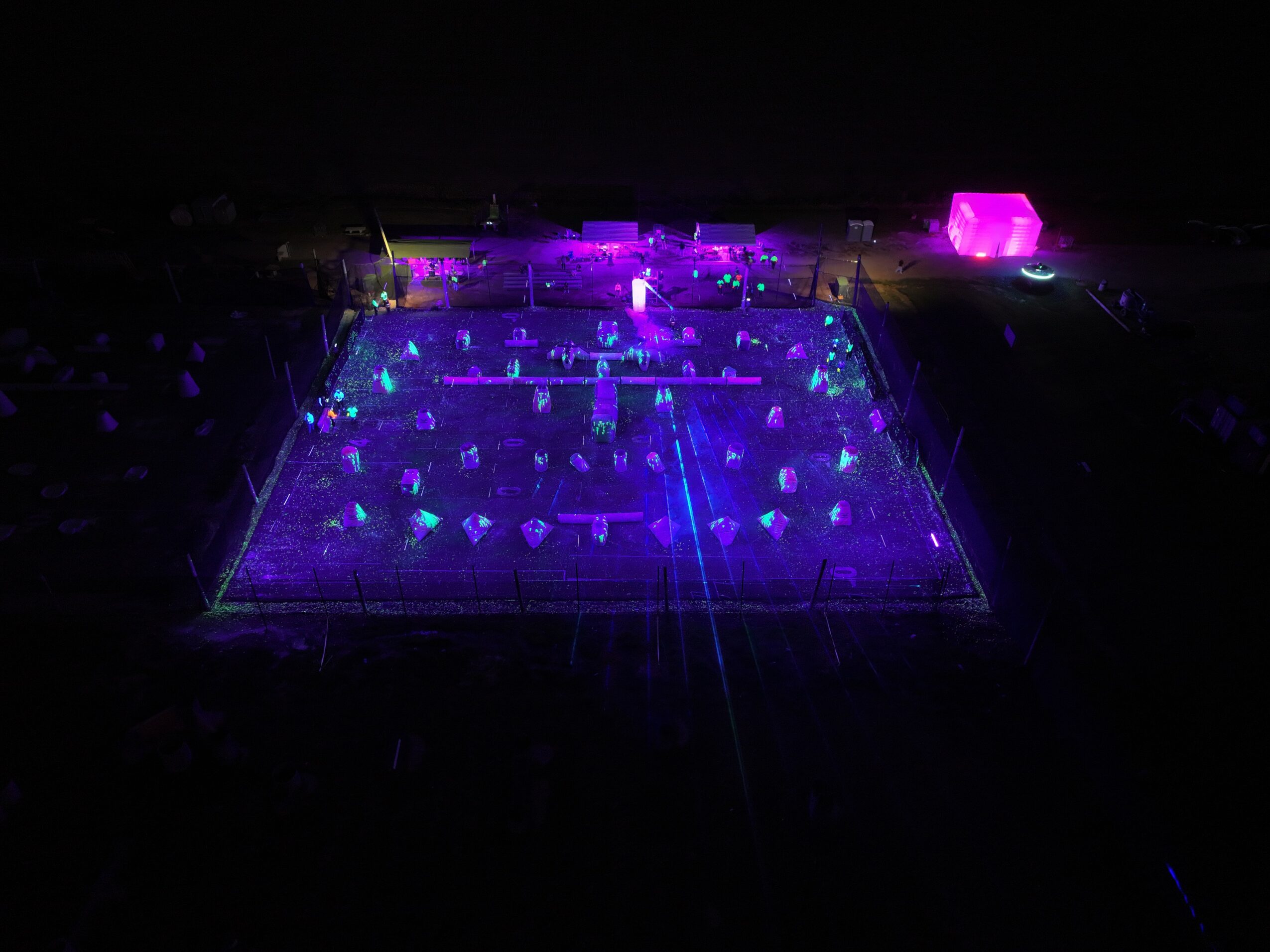 A tennis court illuminated with purple and blue lights at night, with visible net and surrounding area, viewed from above.