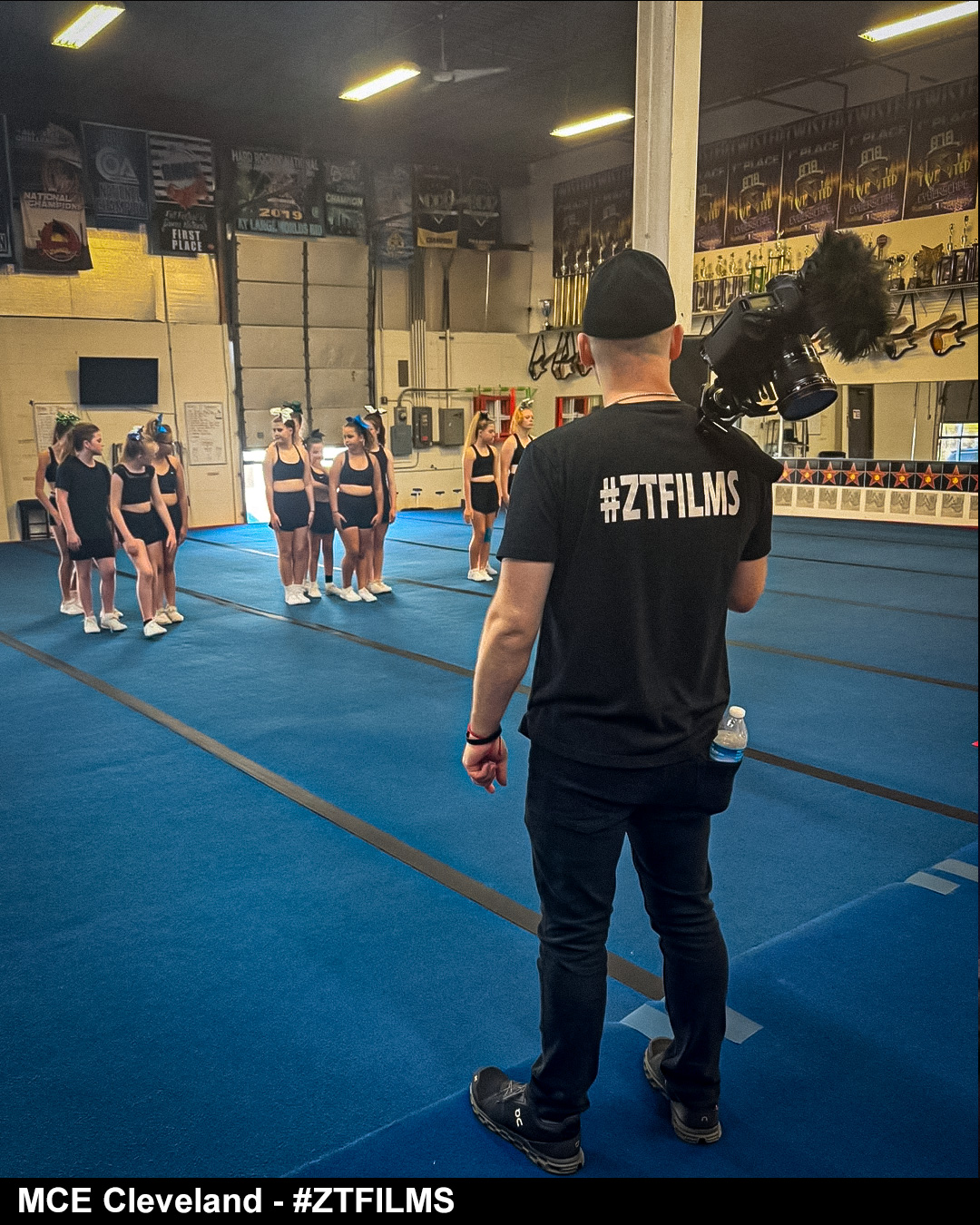 Person filming cheerleaders practicing in a gym with blue mats and banners on the walls.