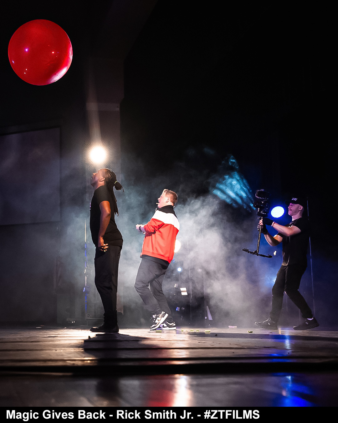 Three performers on stage with smoke, one holding a bright light, and a red ball hanging above. Stage lights illuminate the scene.