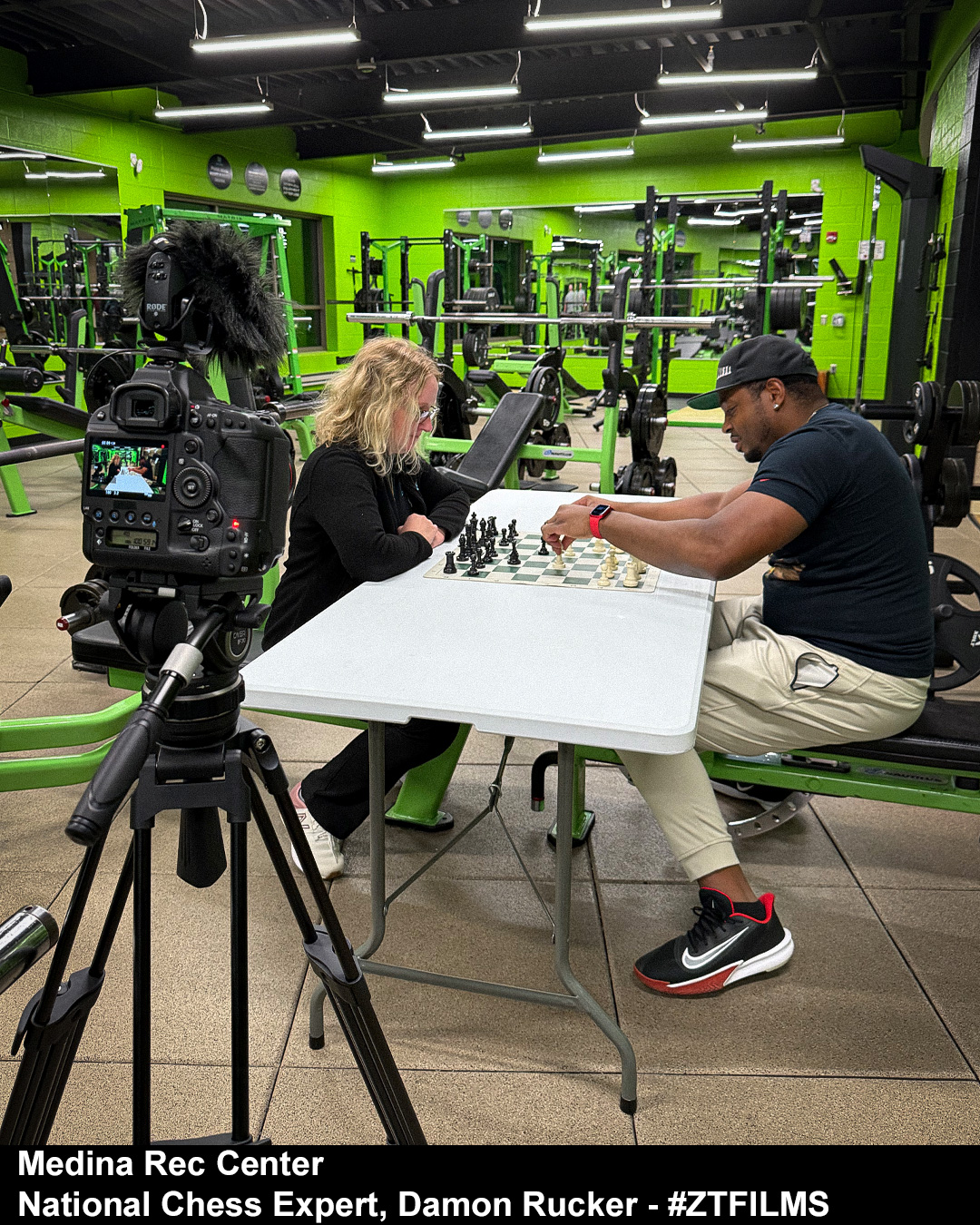 Two people playing chess at a table in a gym with green walls and exercise equipment in the background.
