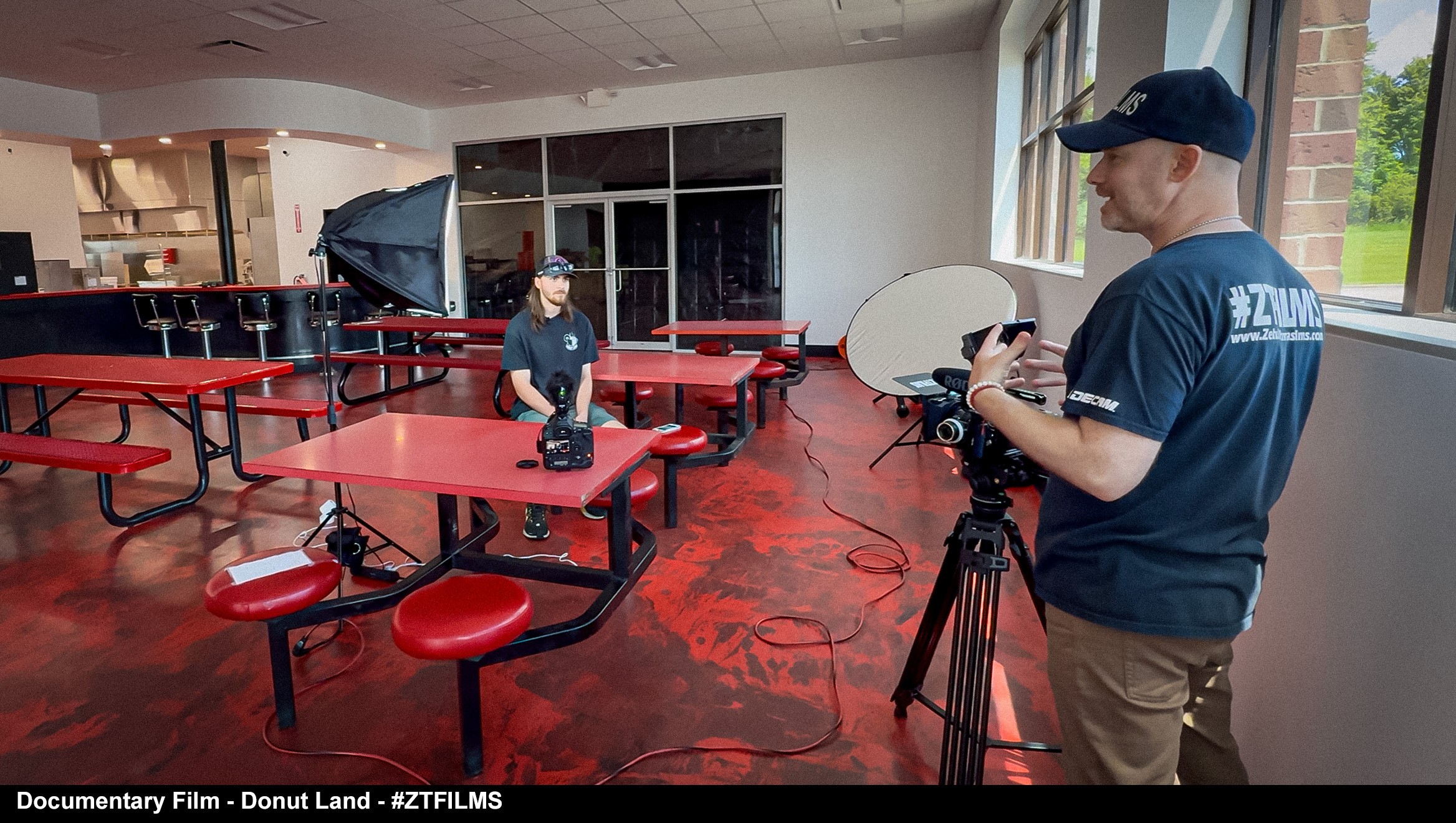 Two people setting up filming equipment in an empty cafeteria with red tables and windows, one person adjusting a camera, the other standing nearby.