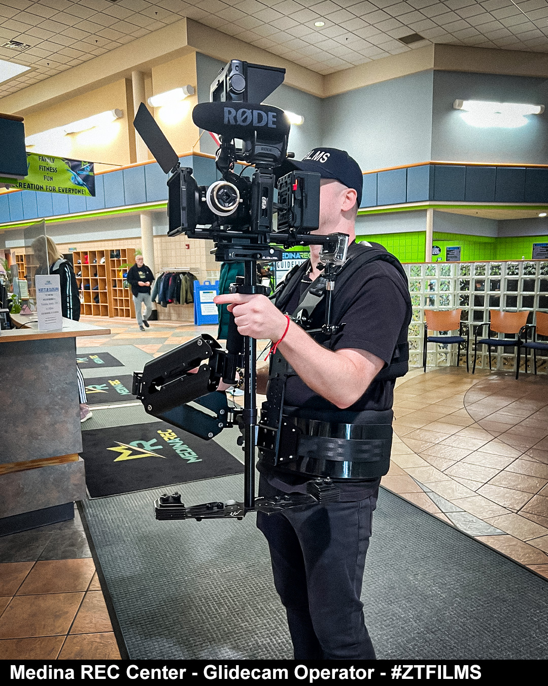 Glidecam operator with camera rig in indoor public space, wearing black clothing and cap, holding equipment with both hands.