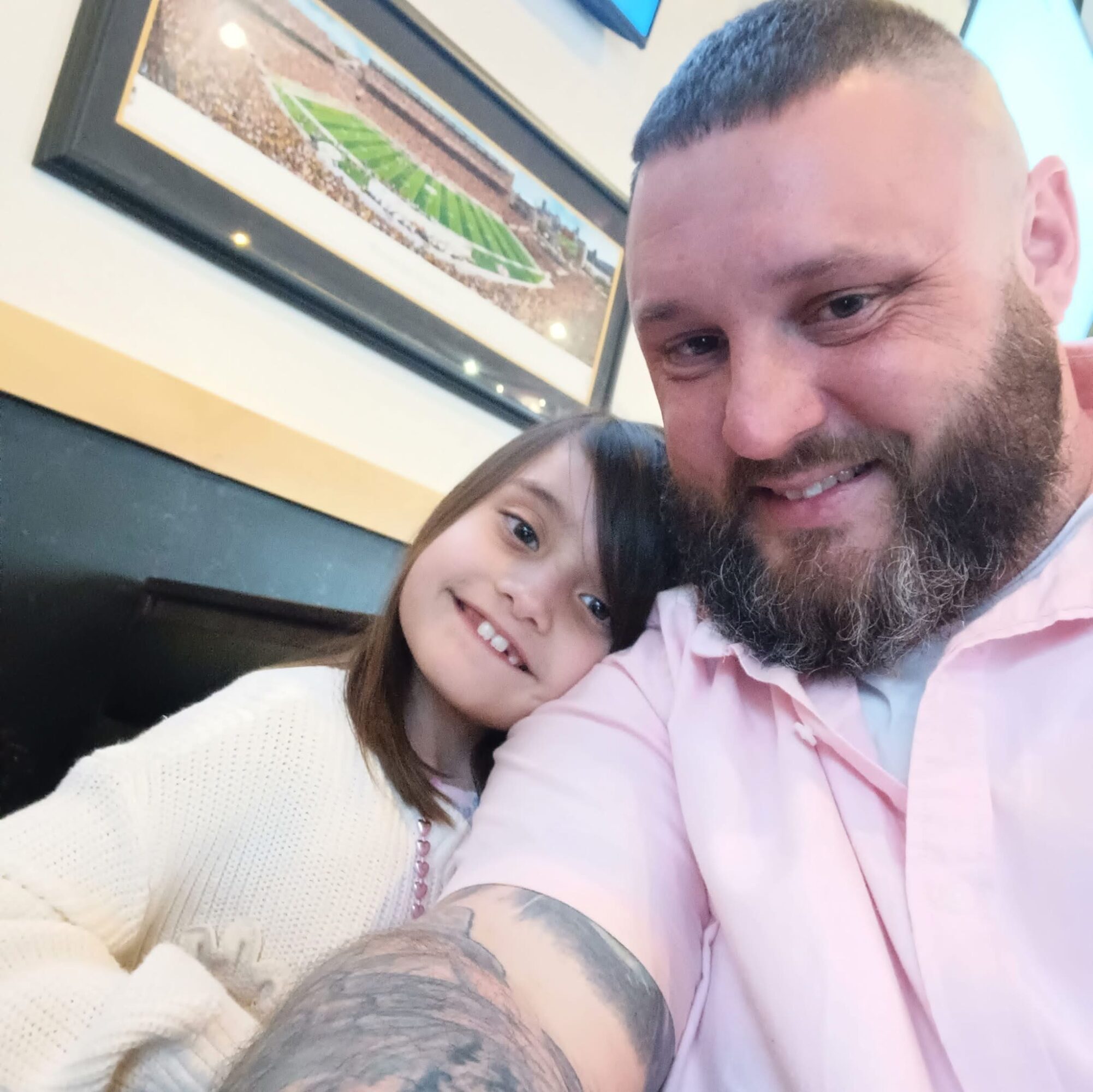 A man and a young girl are smiling and taking a selfie together indoors, with framed pictures on the wall behind them.