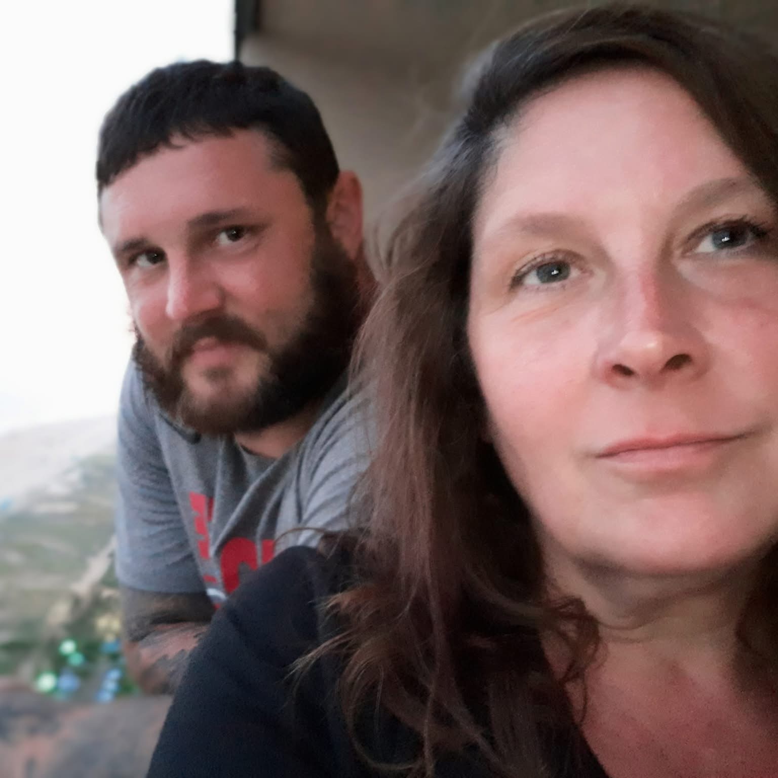 Close-up of a woman with brown hair and a man with a beard, both looking at the camera, outdoors during daytime.
