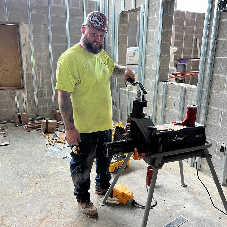 Man in a yellow shirt working with a power tool in a workshop with concrete walls and windows.