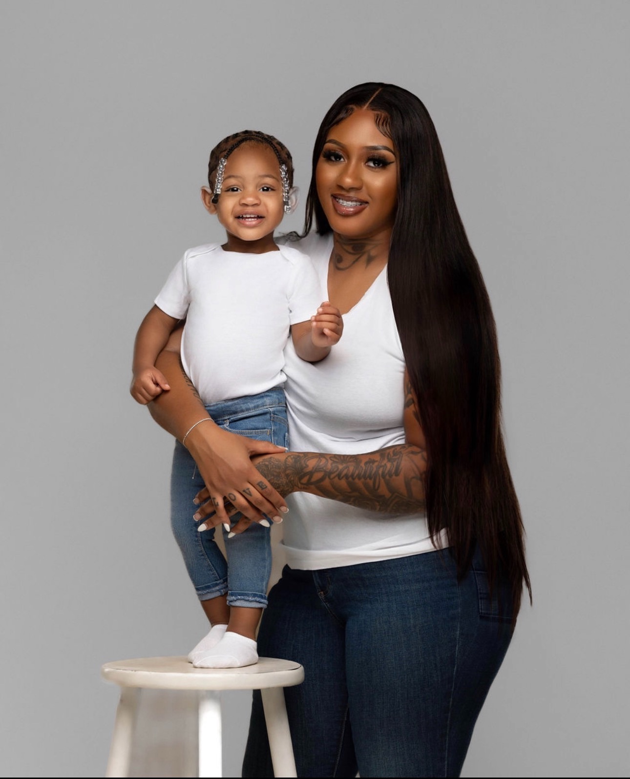 Woman holding a smiling child on a stool, both facing forward, gray background.