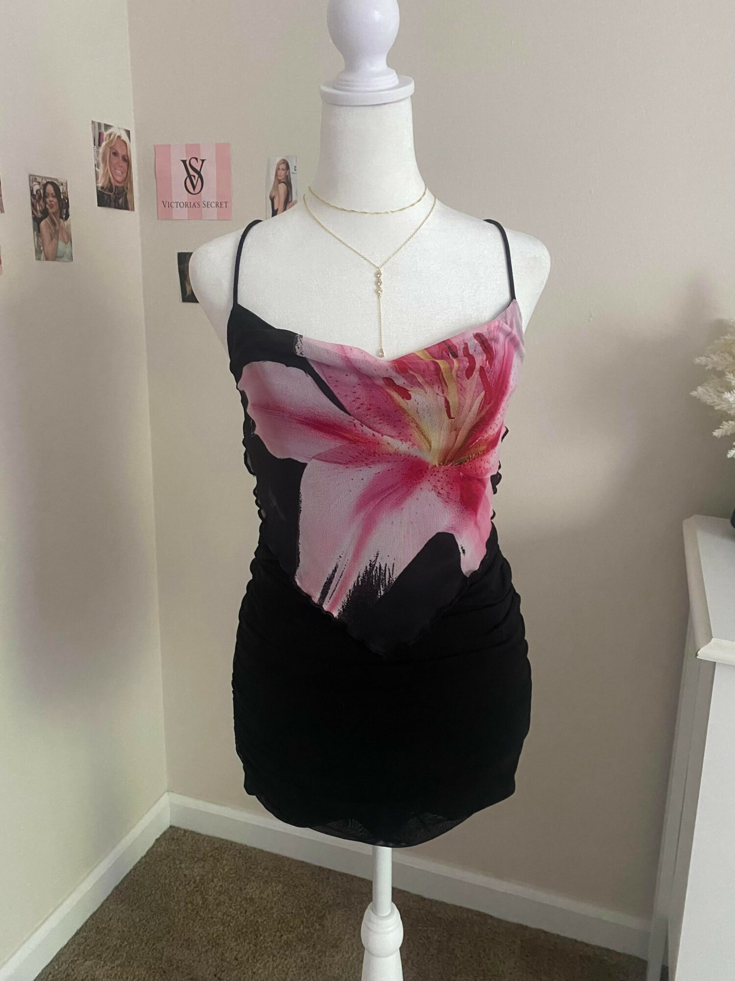Mannequin wearing a black and pink floral dress with spaghetti straps, standing in a room with photos on the wall.