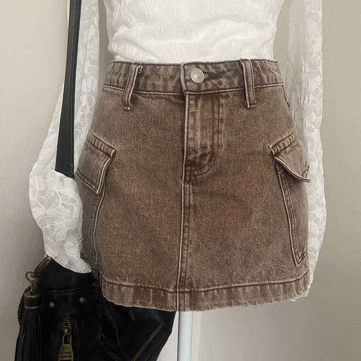 Person wearing a brown denim skirt with pockets, white top, and black bag, standing indoors.
