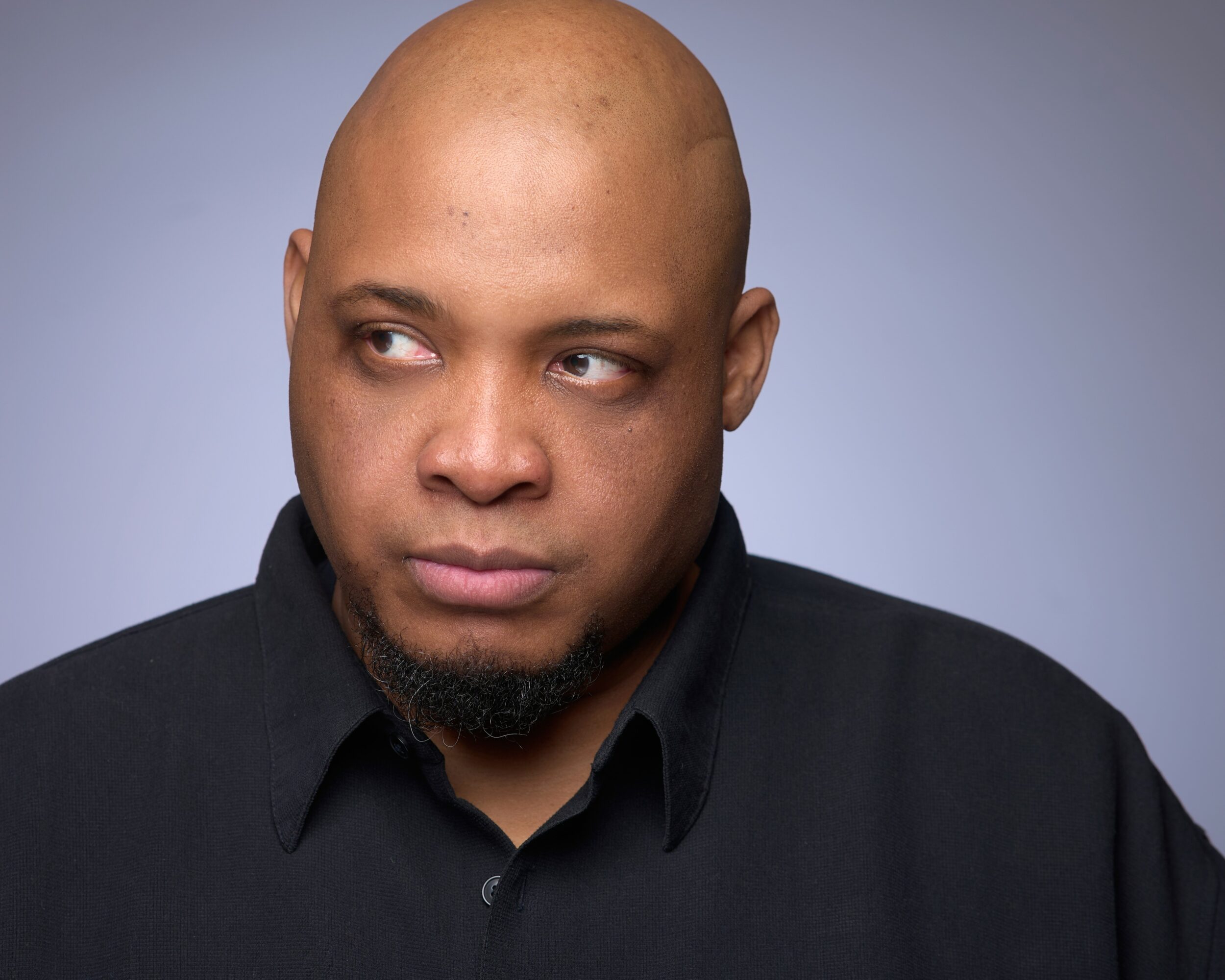 Bald man with a goatee looking to the side, wearing a black shirt, against a plain background.