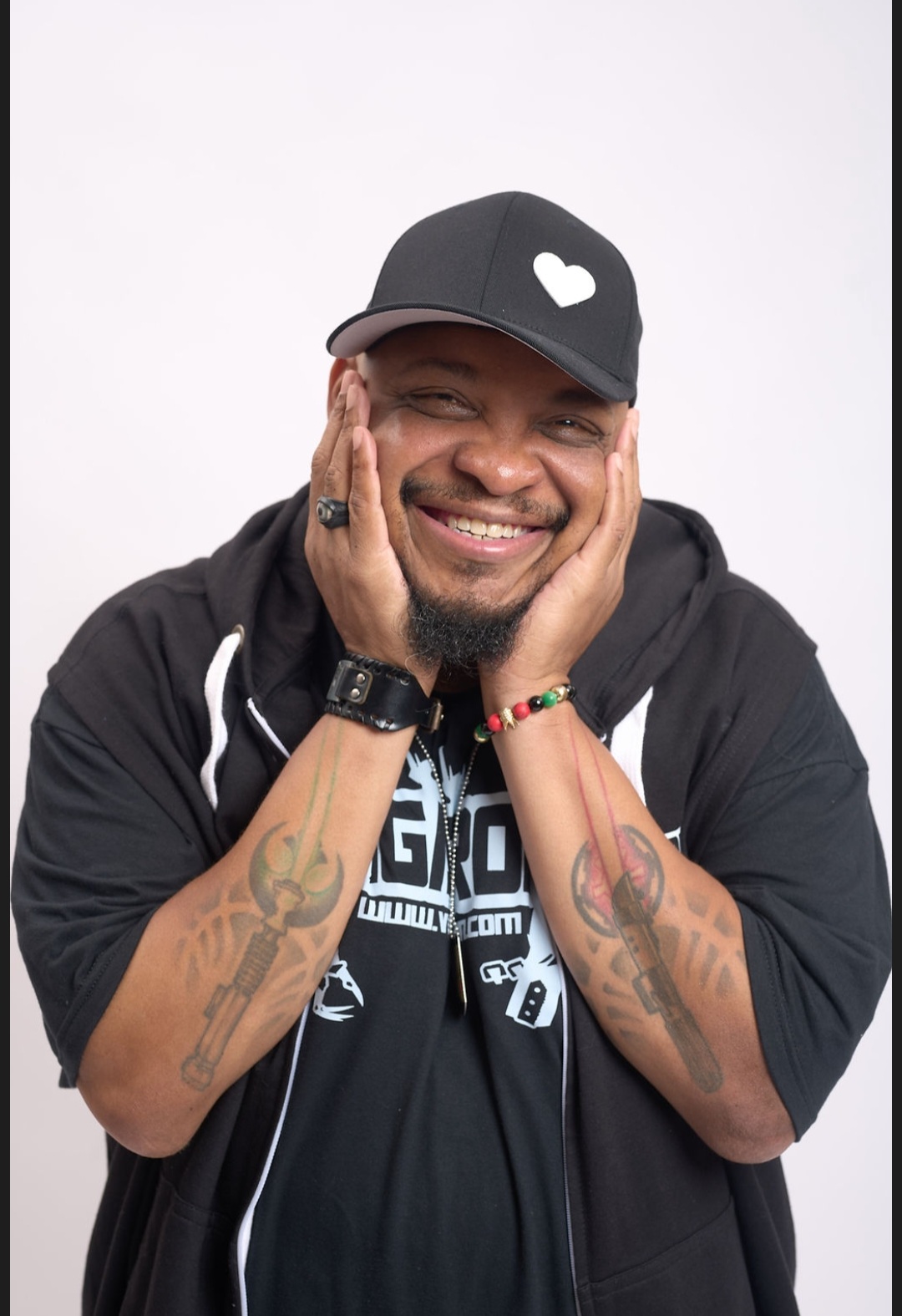 Smiling man with tattoos, wearing a black cap with a heart, black hoodie, and accessories, holding his face with both hands.