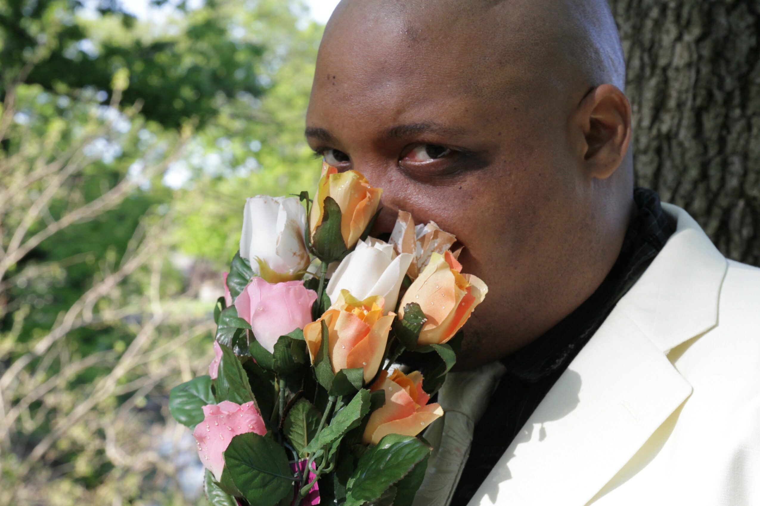 Person with shaved head holding a bouquet of pink, white, and orange roses close to face outdoors.