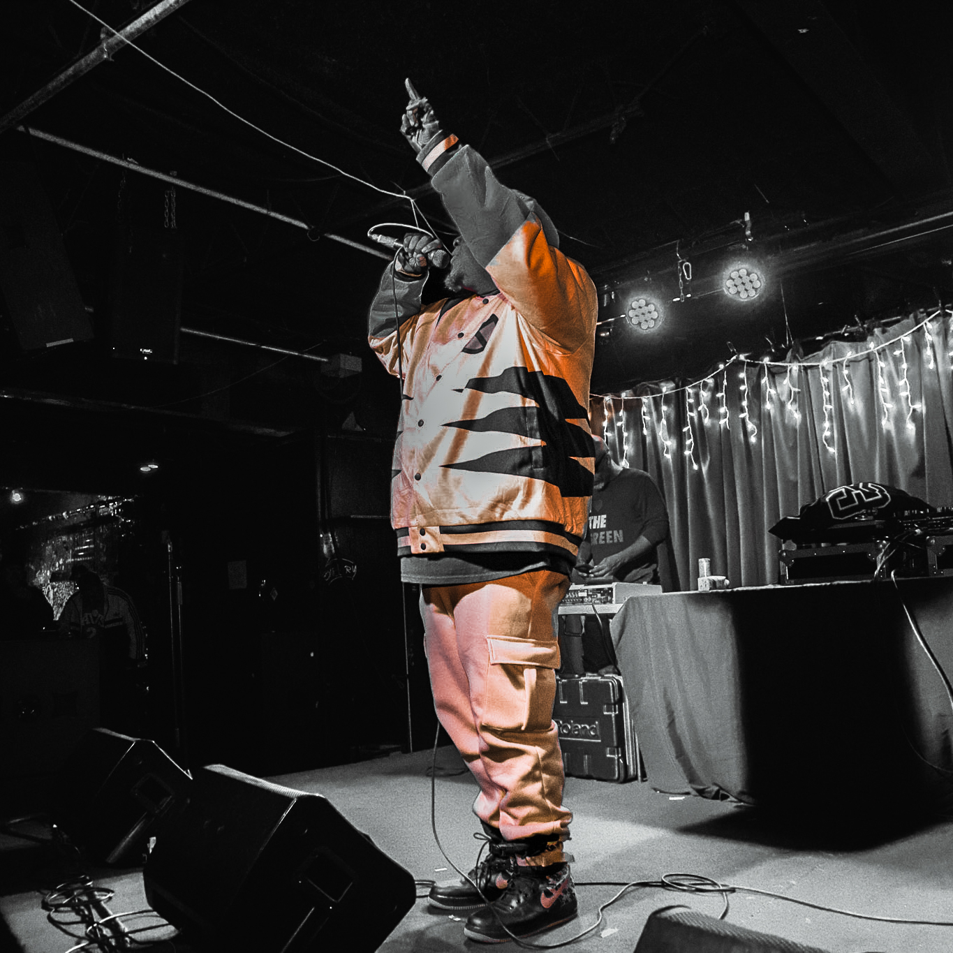 Performer on stage with one arm raised, wearing a jacket and pants, in a dimly lit venue with string lights.