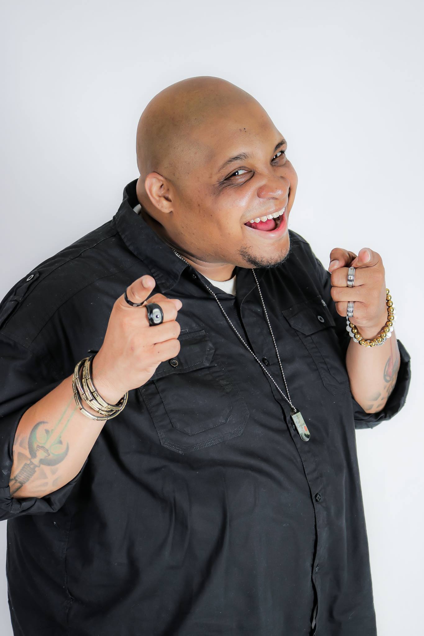 Person with a shaved head smiling, wearing a black shirt, jewelry, and making a fist gesture, against a white background.