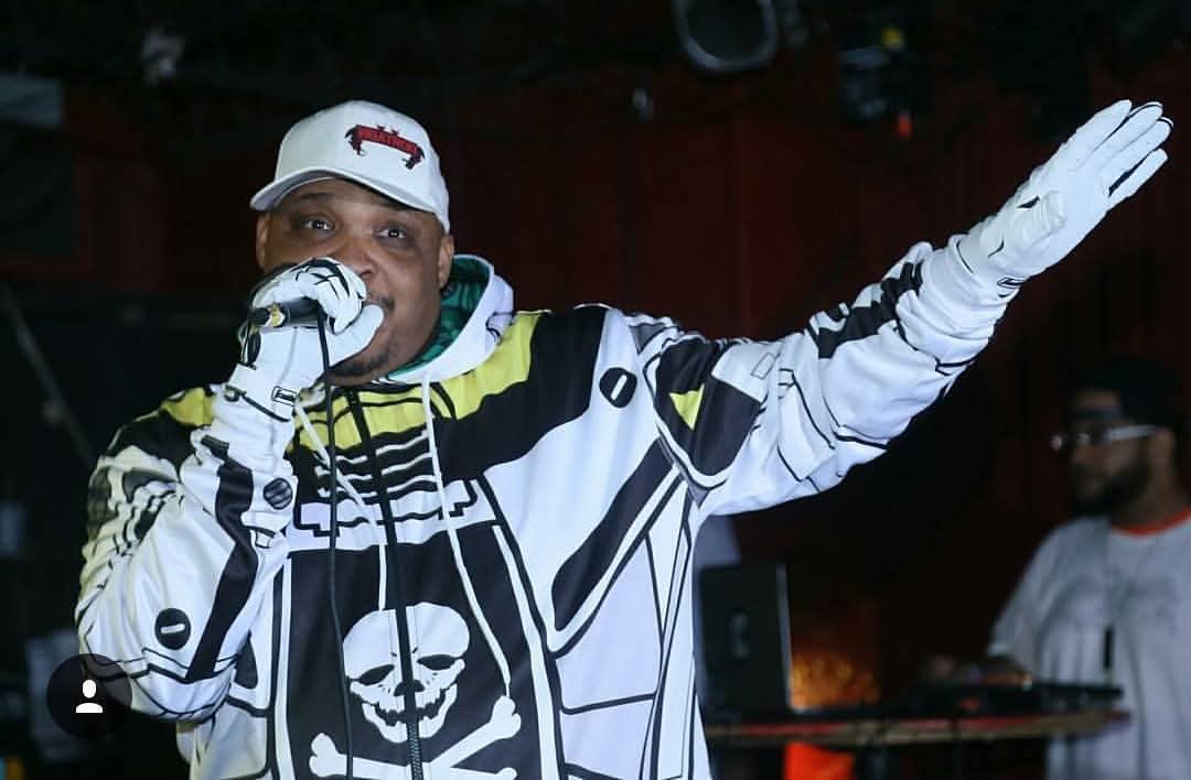 Performer wearing a skull-patterned jacket and white cap, singing into a microphone with one arm raised, stage background visible.