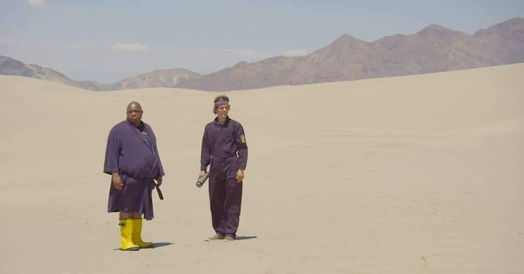 Two people walking in a desert with sand dunes and mountains in the background.