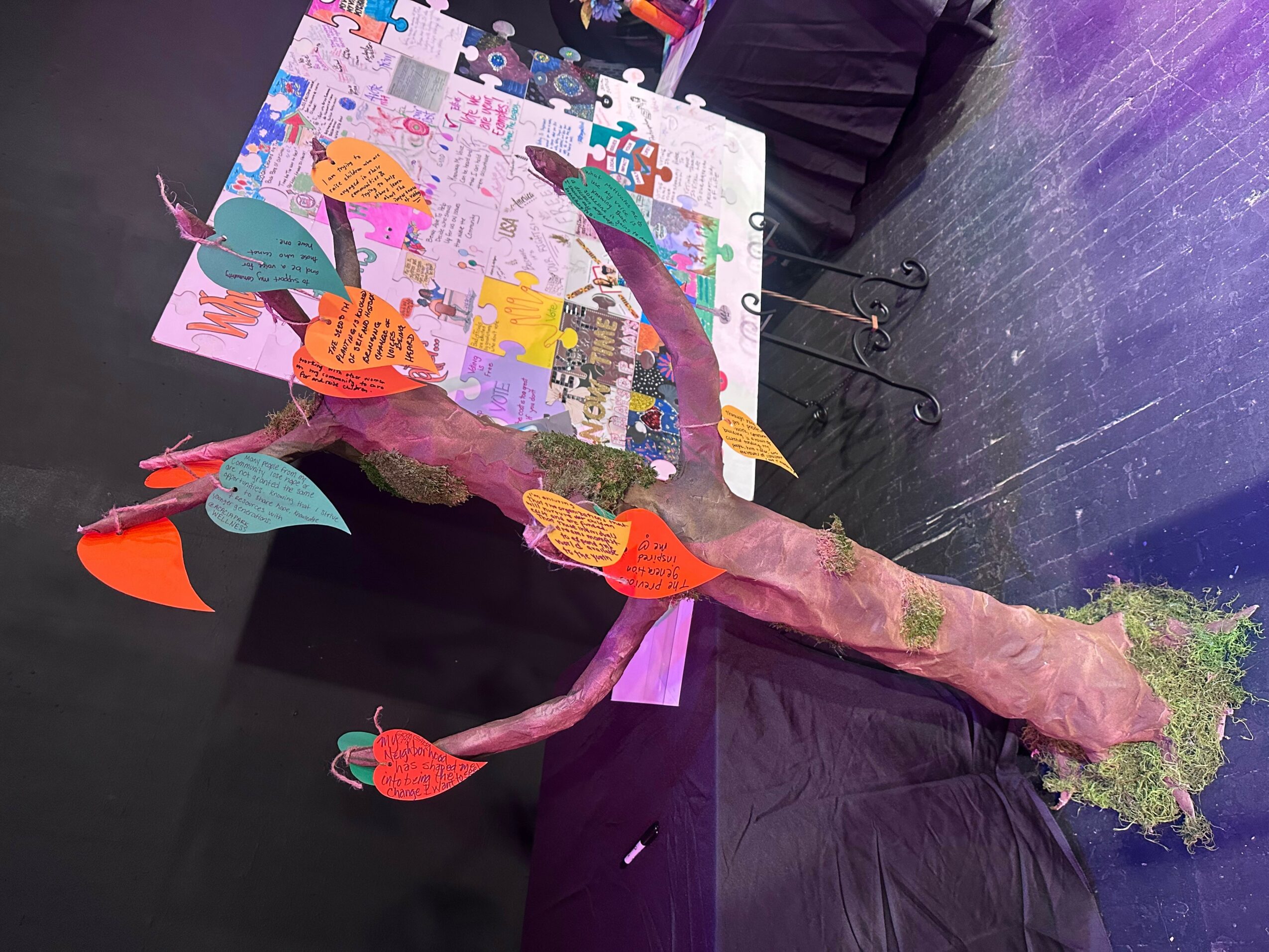 Colorful paper fish hanging from a tree branch, with a display of drawings or posters in the background.