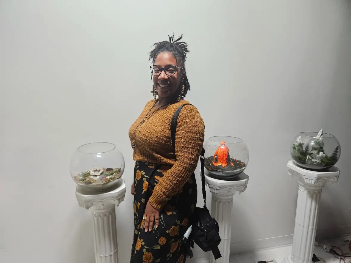 Person with glasses and dreadlocks smiling, standing between three glass terrariums on white pedestals, against a plain white wall.