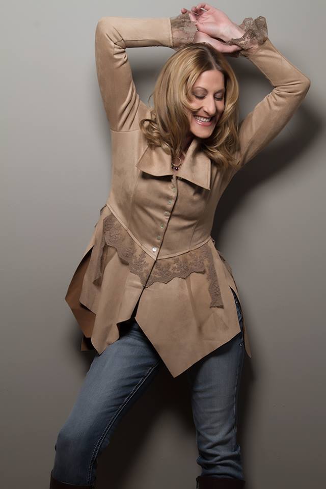 Woman with blonde hair smiling, arms raised, wearing a beige coat and jeans, standing against a plain wall.