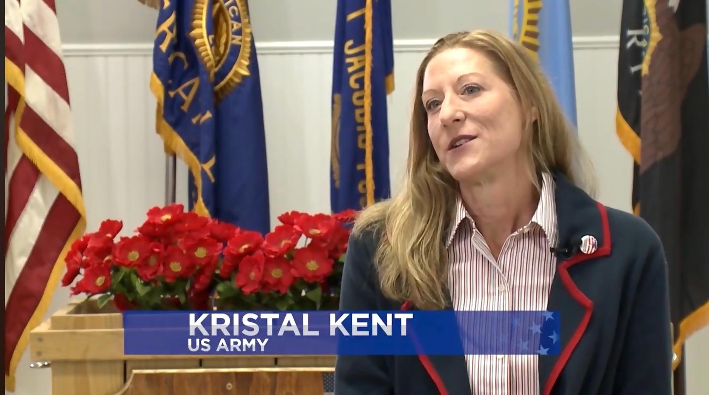 Woman with long blonde hair speaking in front of flags and red flowers, with a name tag reading 'Kristal Kent, US Army'.
