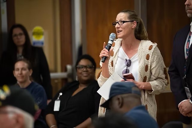 Woman speaking into microphone at indoor event, holding papers, wearing glasses and a beige cardigan with cutouts, smiling.