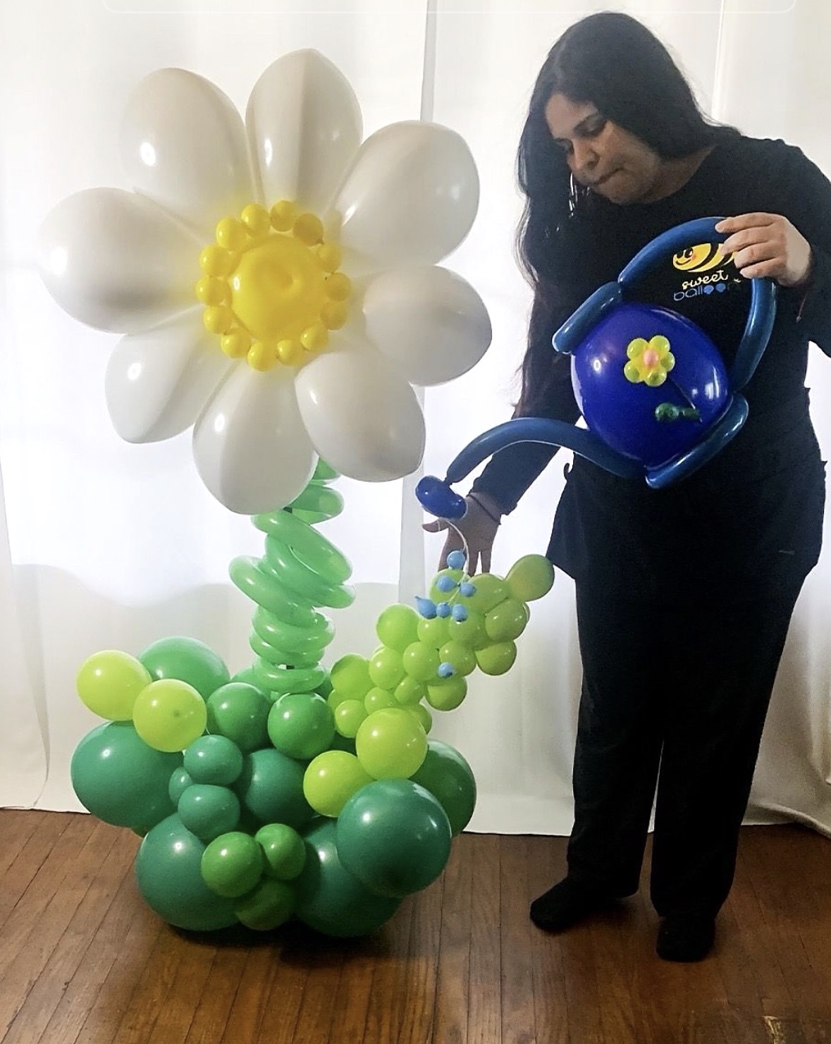 Woman watering a balloon flower sculpture with white petals, yellow center, green stem, and leaves, indoors with white curtains behind.