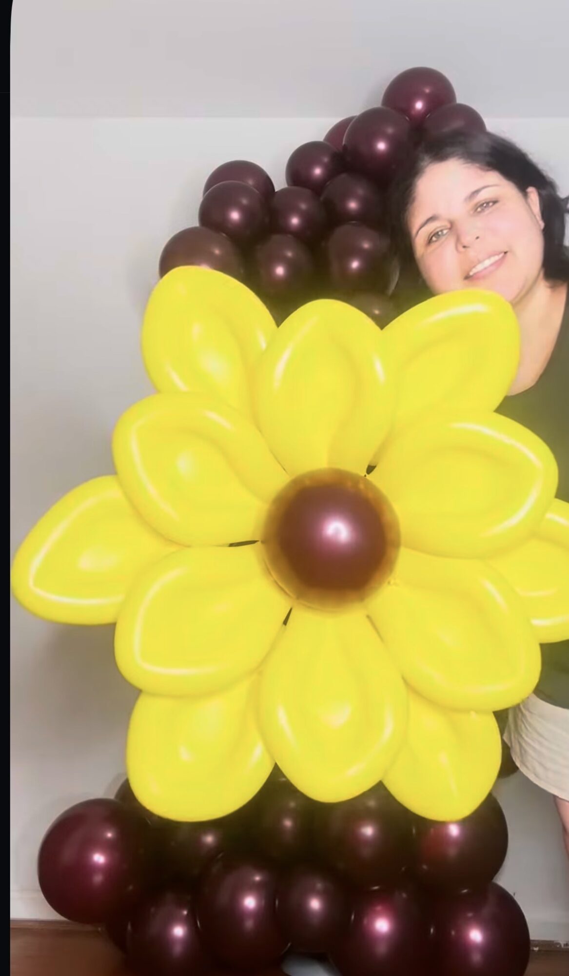 Person with dark hair smiling behind a large yellow flower-shaped balloon arrangement with purple balloons in the background.