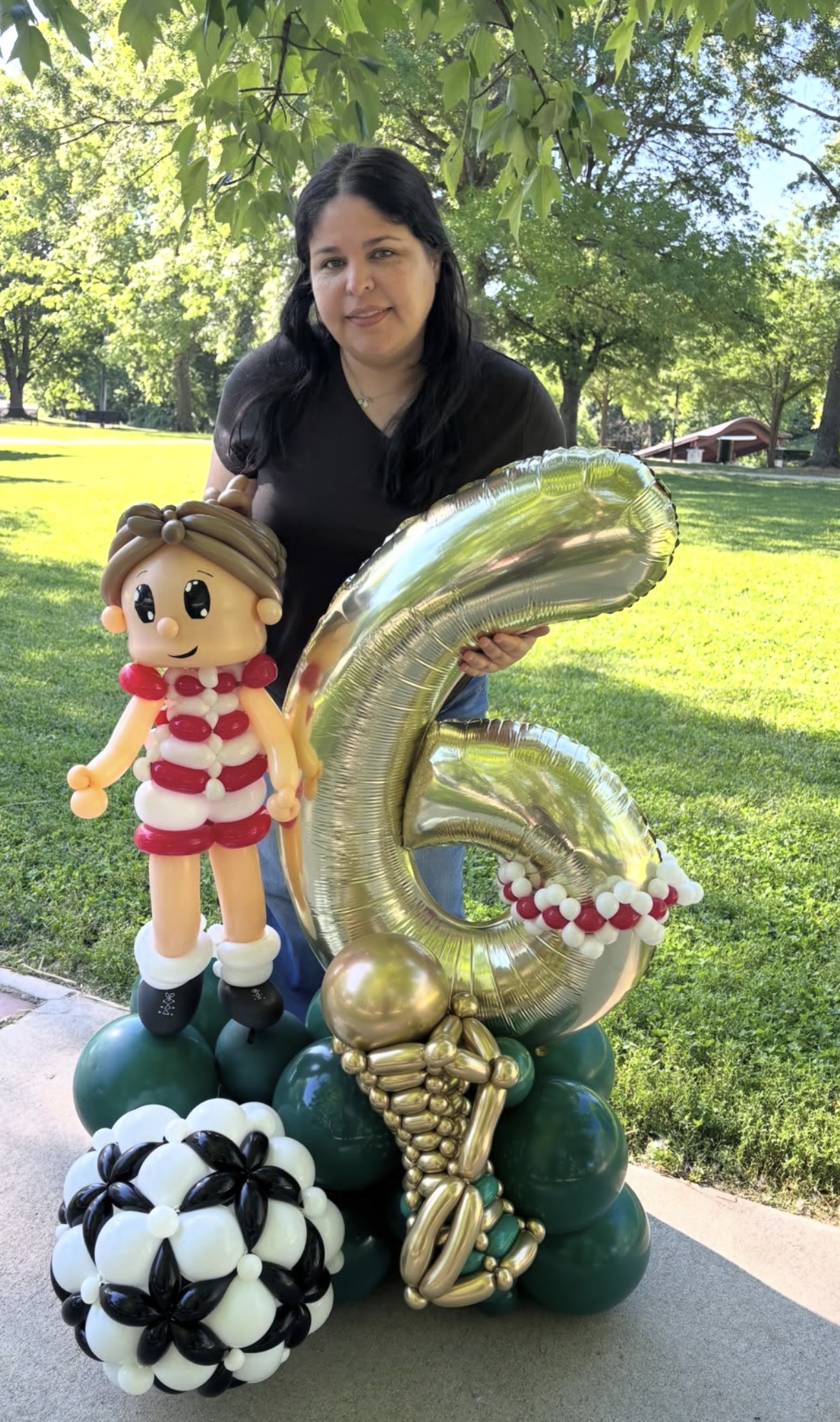 Woman holding a large gold number six balloon, with a soccer ball, a doll, and a bouquet of flowers, outdoors in a park.