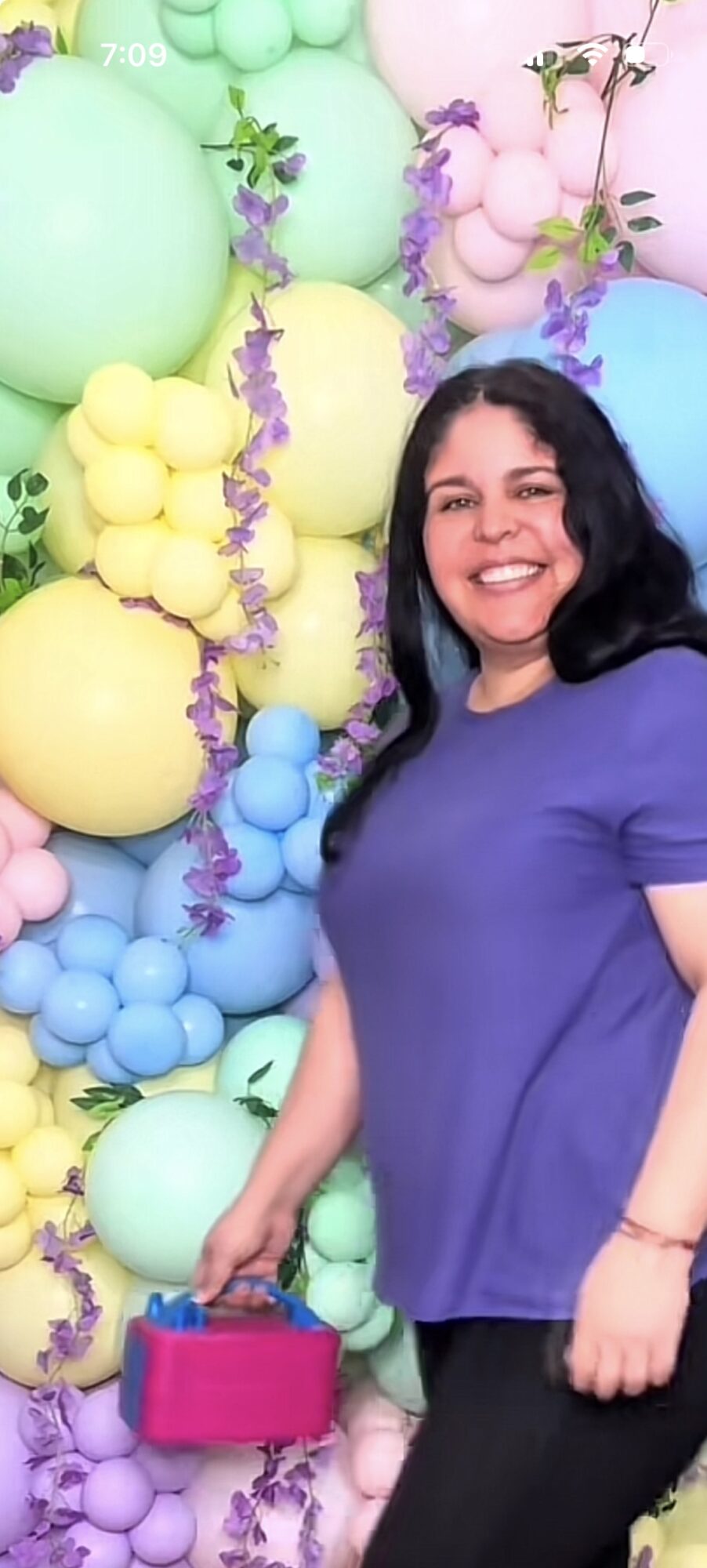 Woman smiling in front of colorful balloons, holding a pink basket, wearing a blue shirt and black pants.