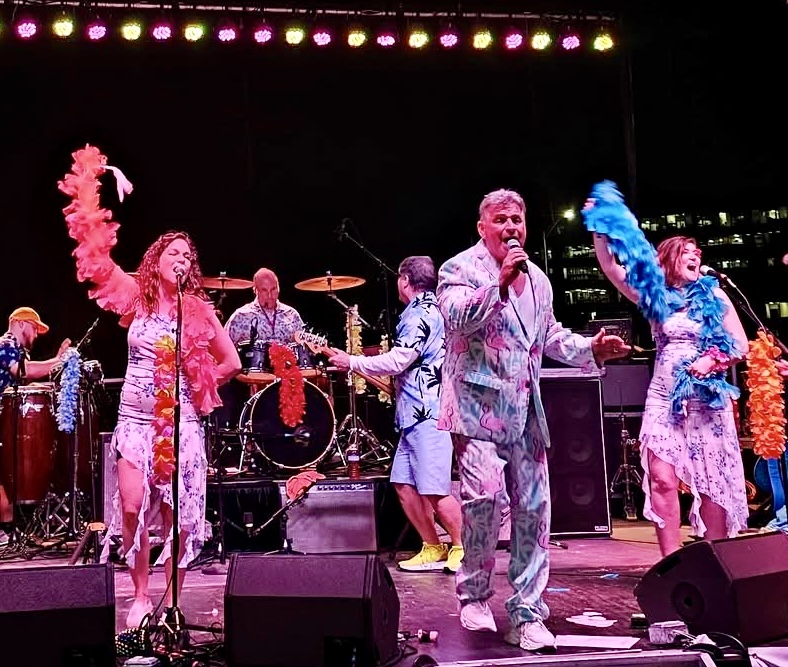 Group of performers on stage singing and dancing with colorful costumes and musical instruments, stage lights overhead.