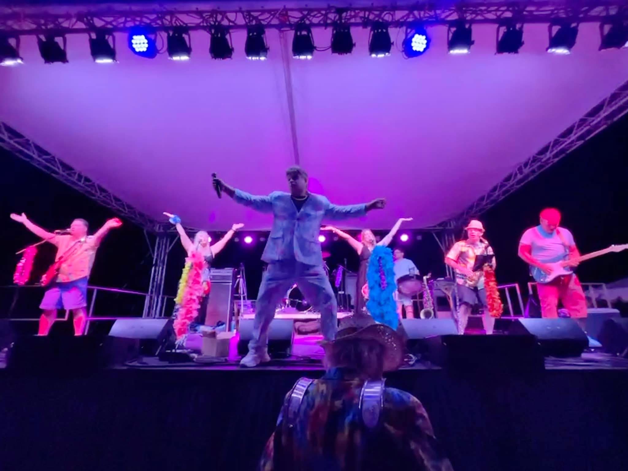 Group of six people performing on stage with a singer in the center, colorful lighting, and musical instruments in the background.