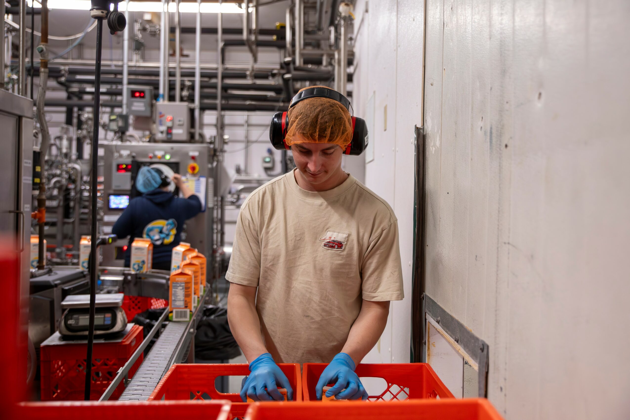 Young worker in beige shirt and blue gloves operating machinery in industrial setting, with another worker in background.