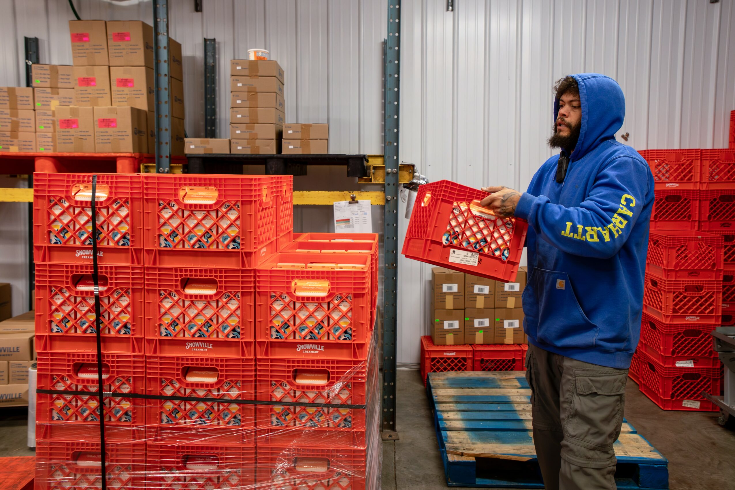Man in blue hoodie holding a red shopping basket in a warehouse with stacked red crates and cardboard boxes