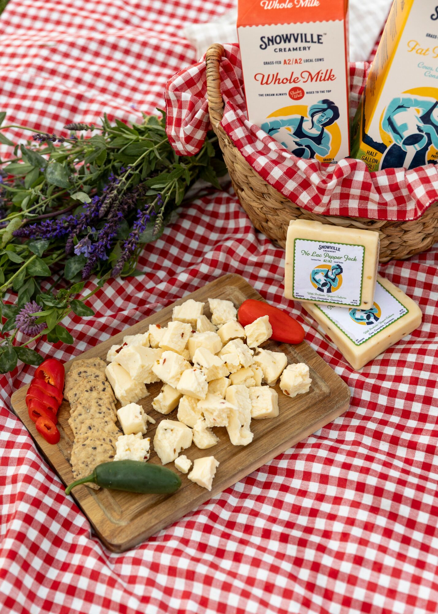 Cheese on a wooden board with red chili and green chili peppers, lavender flowers, and a basket of milk chocolate bars on a red and white checkered cloth.