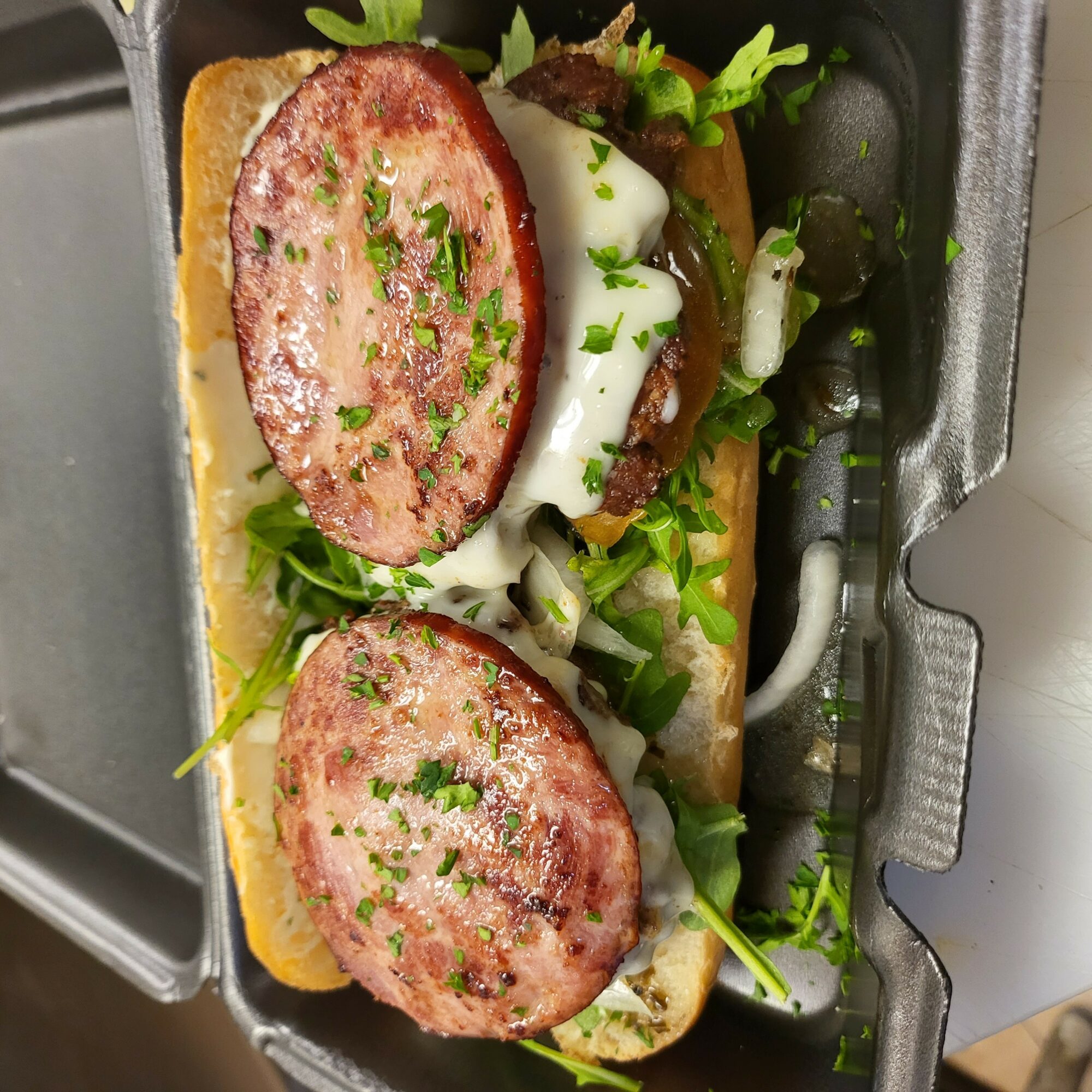 Sandwich with sausage slices, lettuce, and sauce in a container, viewed from the side.