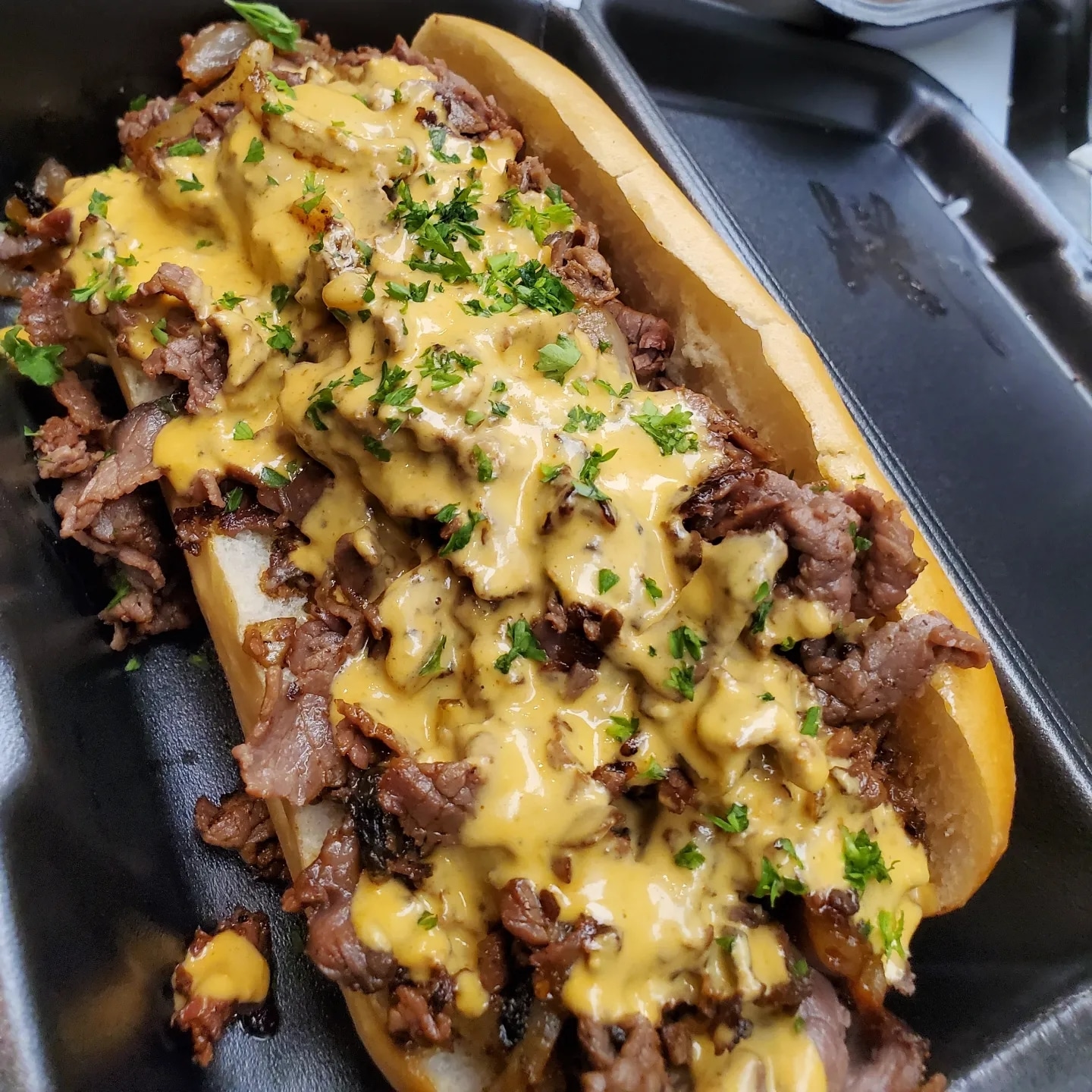Hot beef and cheese sandwich with chopped herbs on a bun, served in a black container.