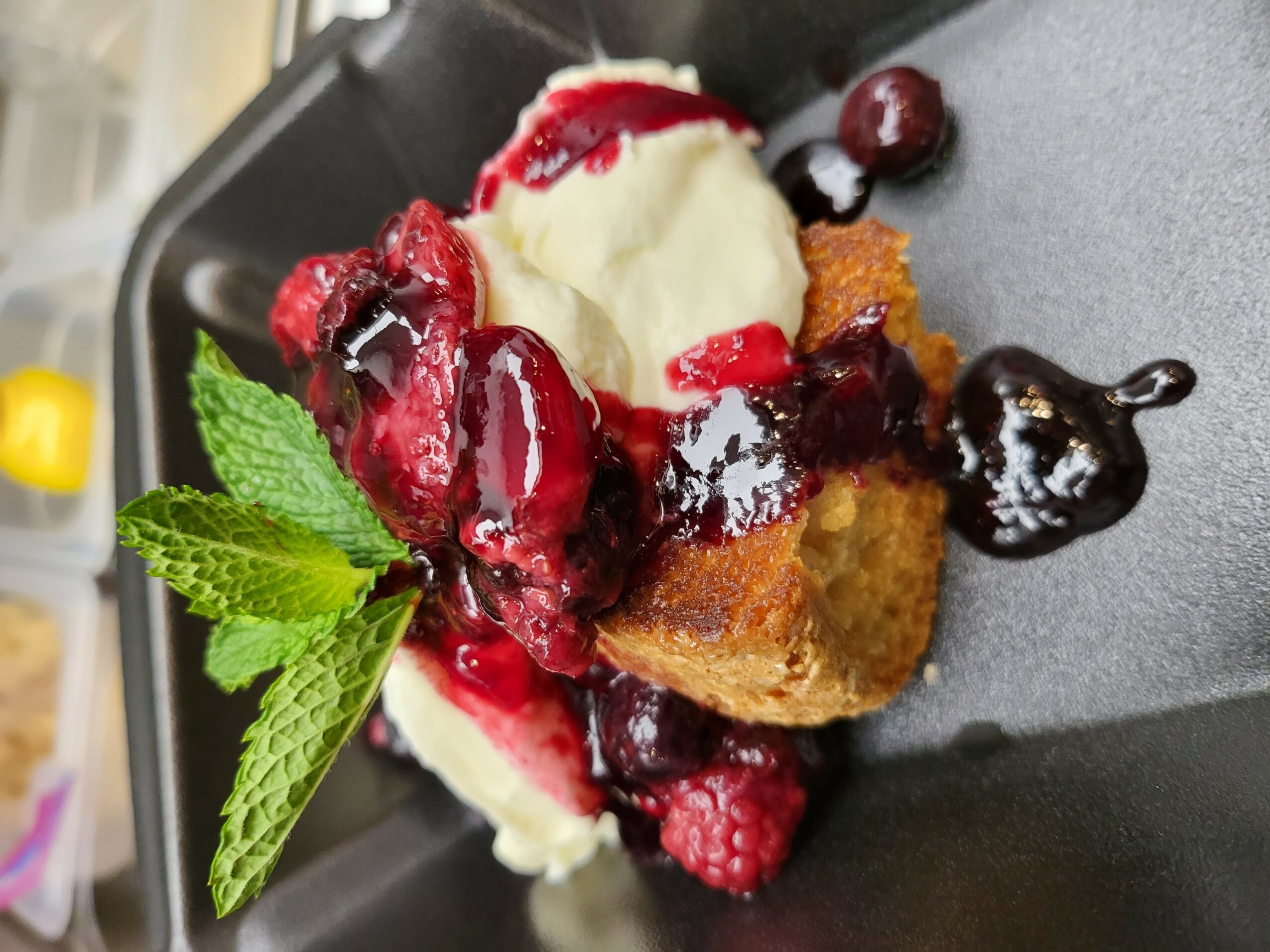 Dessert with ice cream, mixed berries, mint leaves, and sauce on a black plate.