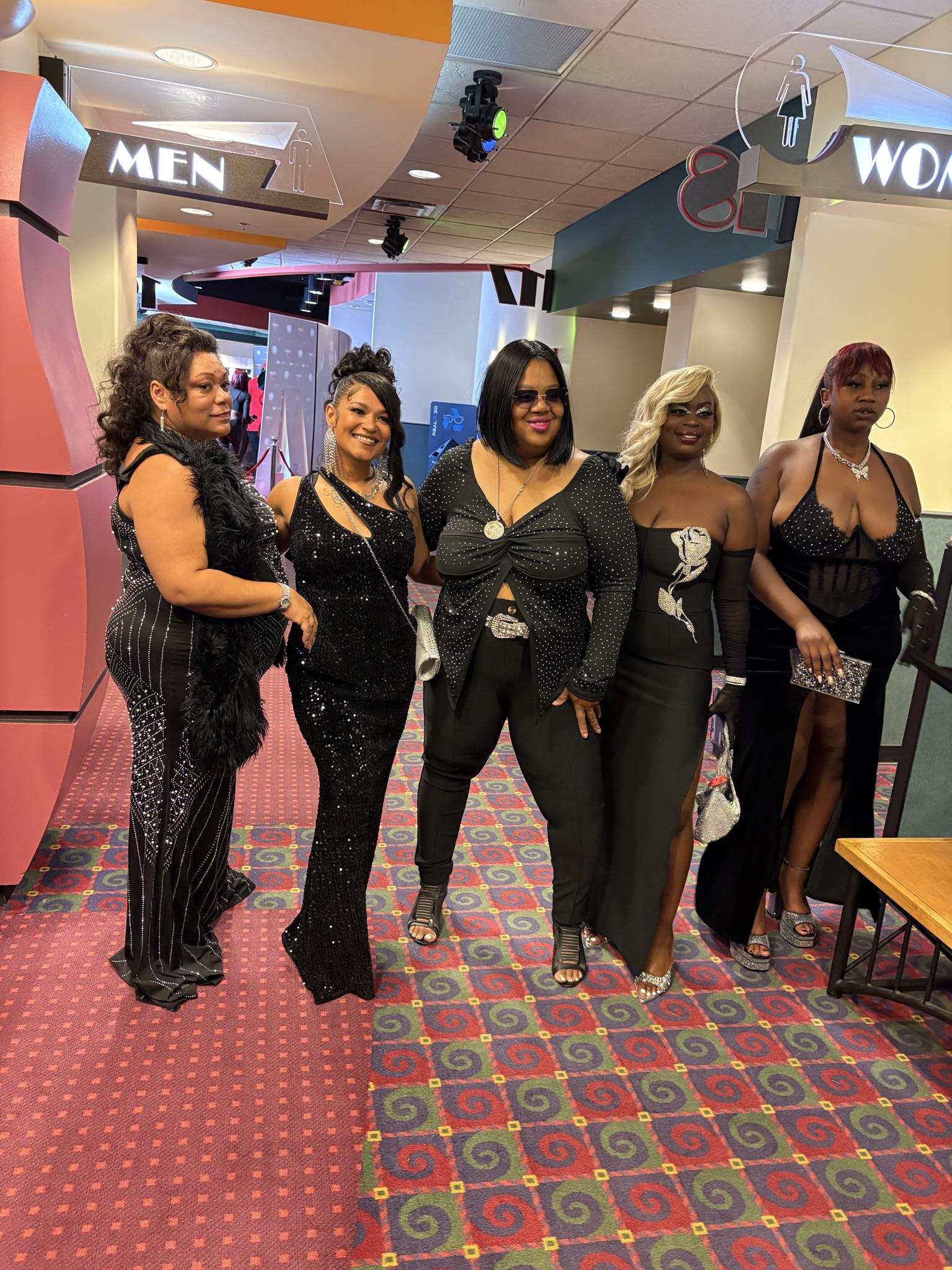 Five women stand together in a shopping mall, dressed in black outfits, smiling, with store signs visible above them.