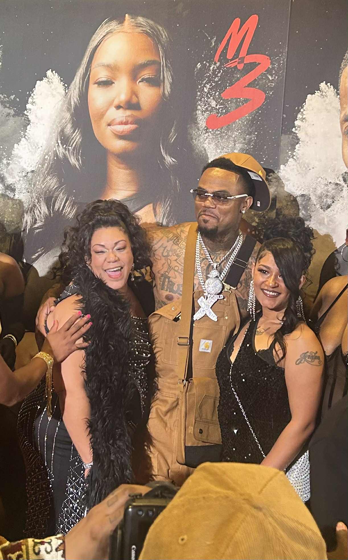 Three people smiling and posing in front of a large poster of a woman's face with the text 'M3'.