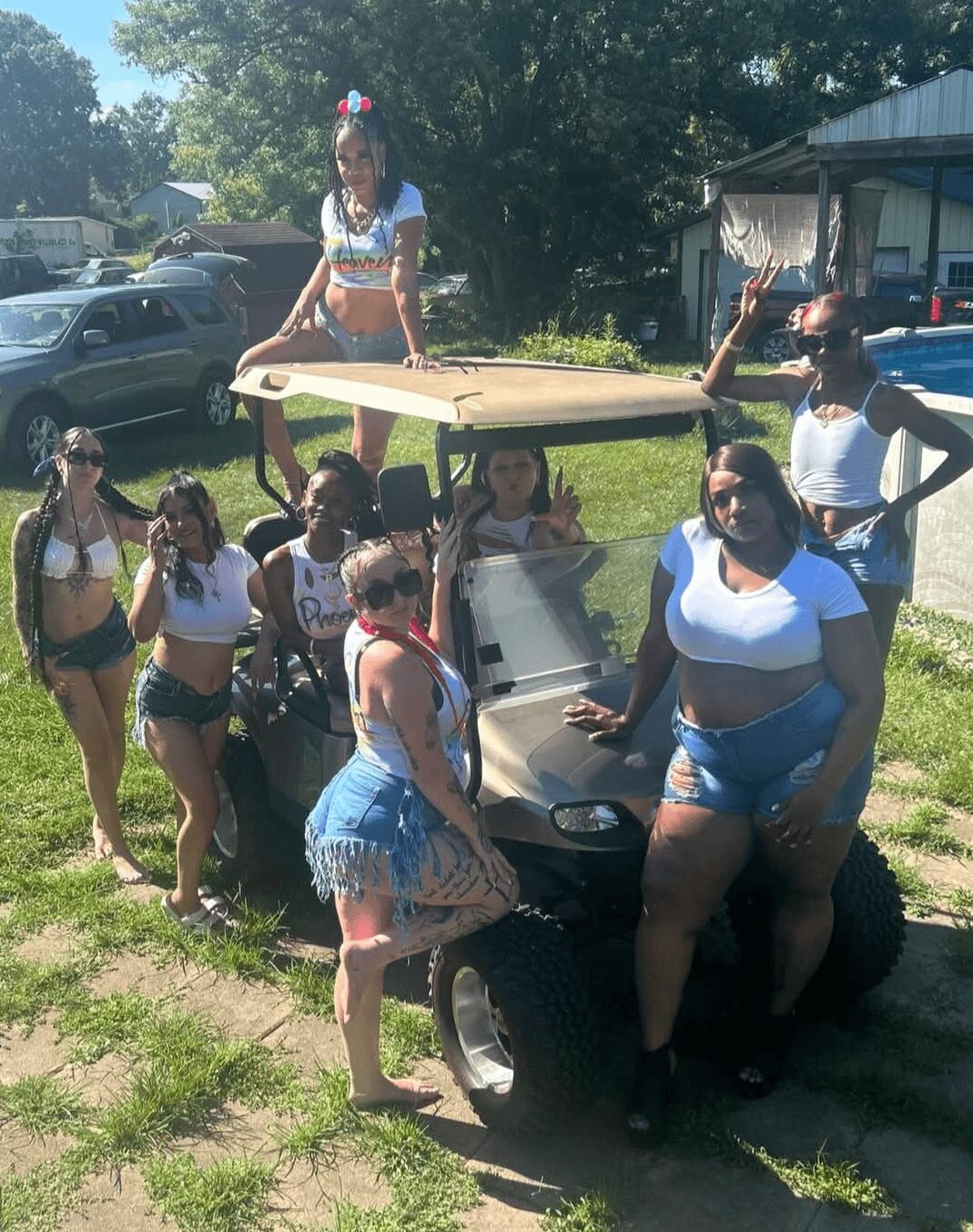 Group of women and children outdoors with a golf cart, some standing and one sitting on the cart, in a grassy area.