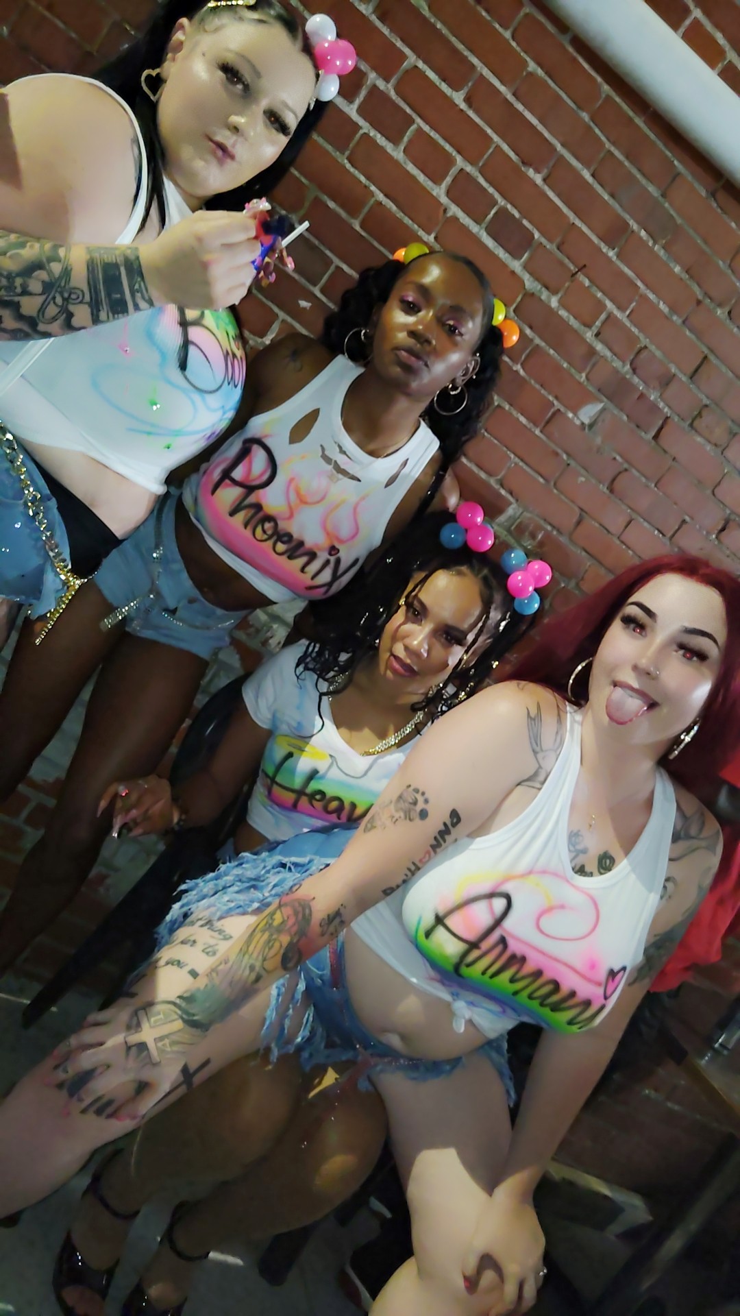 Four young women with colorful accessories and tattoos pose against a brick wall, smiling and making playful gestures.
