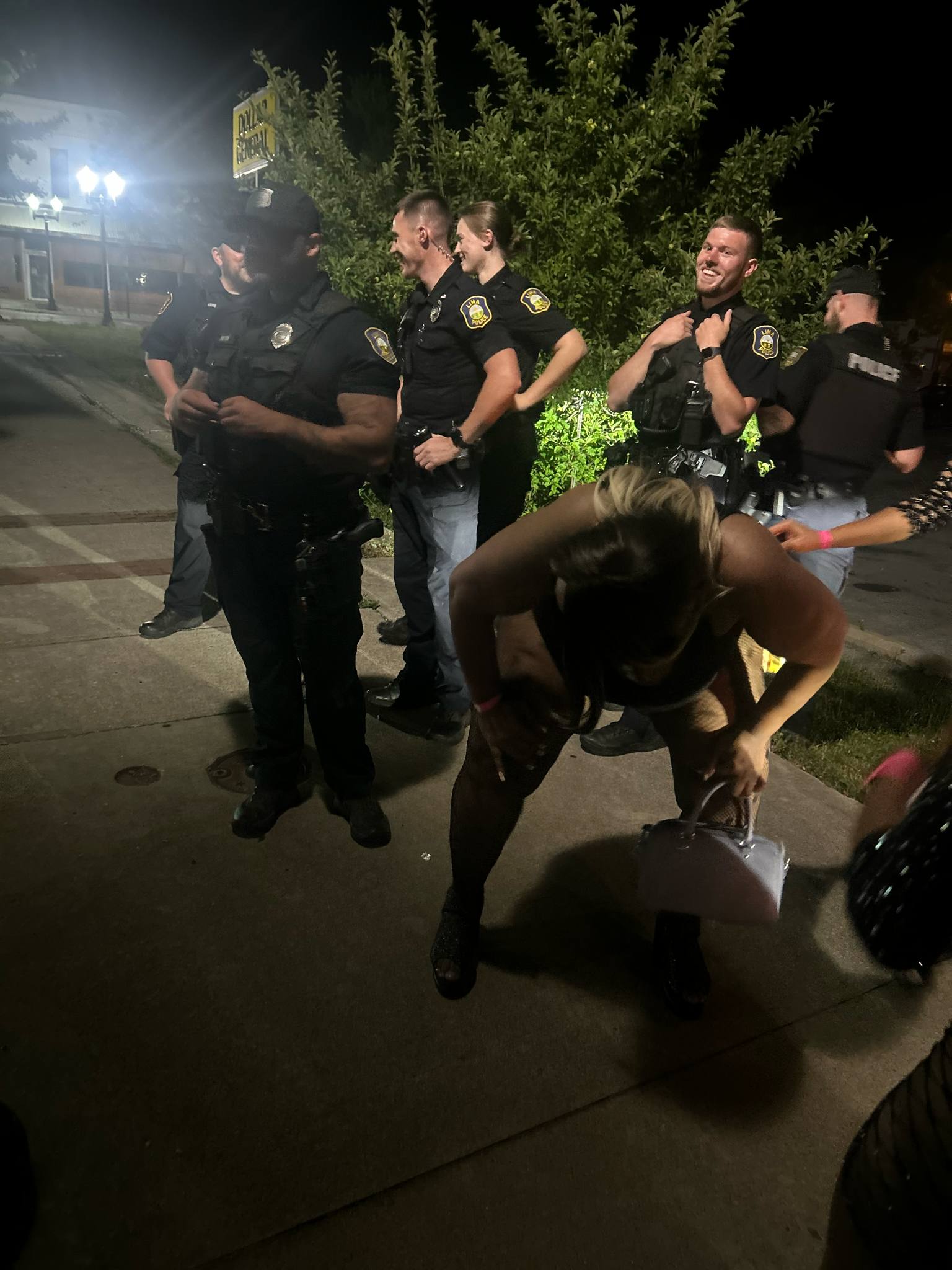 Group of police officers standing outdoors at night, with a woman bending down in front of them, some smiling and others looking serious.