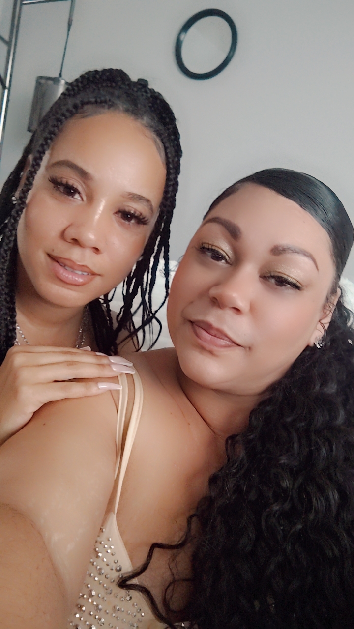 Two women with braided and curly hair pose closely indoors, smiling softly, with a white wall and a small round mirror behind them.