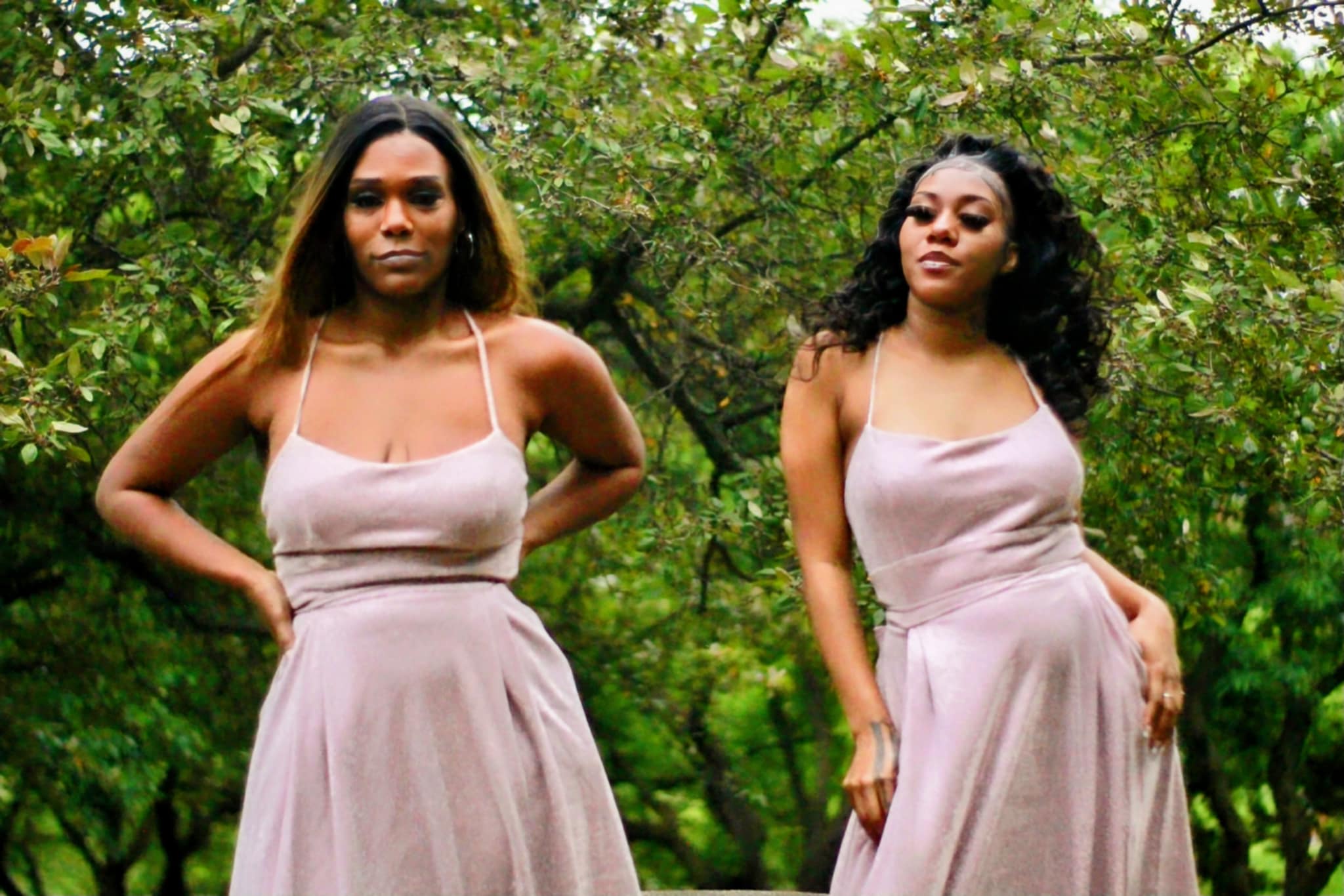 Two women stand outdoors in front of green trees, wearing matching light-colored dresses, with serious expressions.