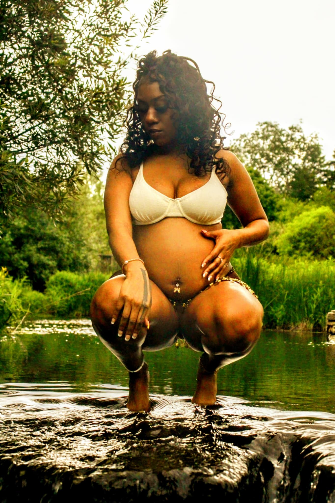 Pregnant woman crouching in water outdoors, holding her belly, surrounded by trees and greenery.