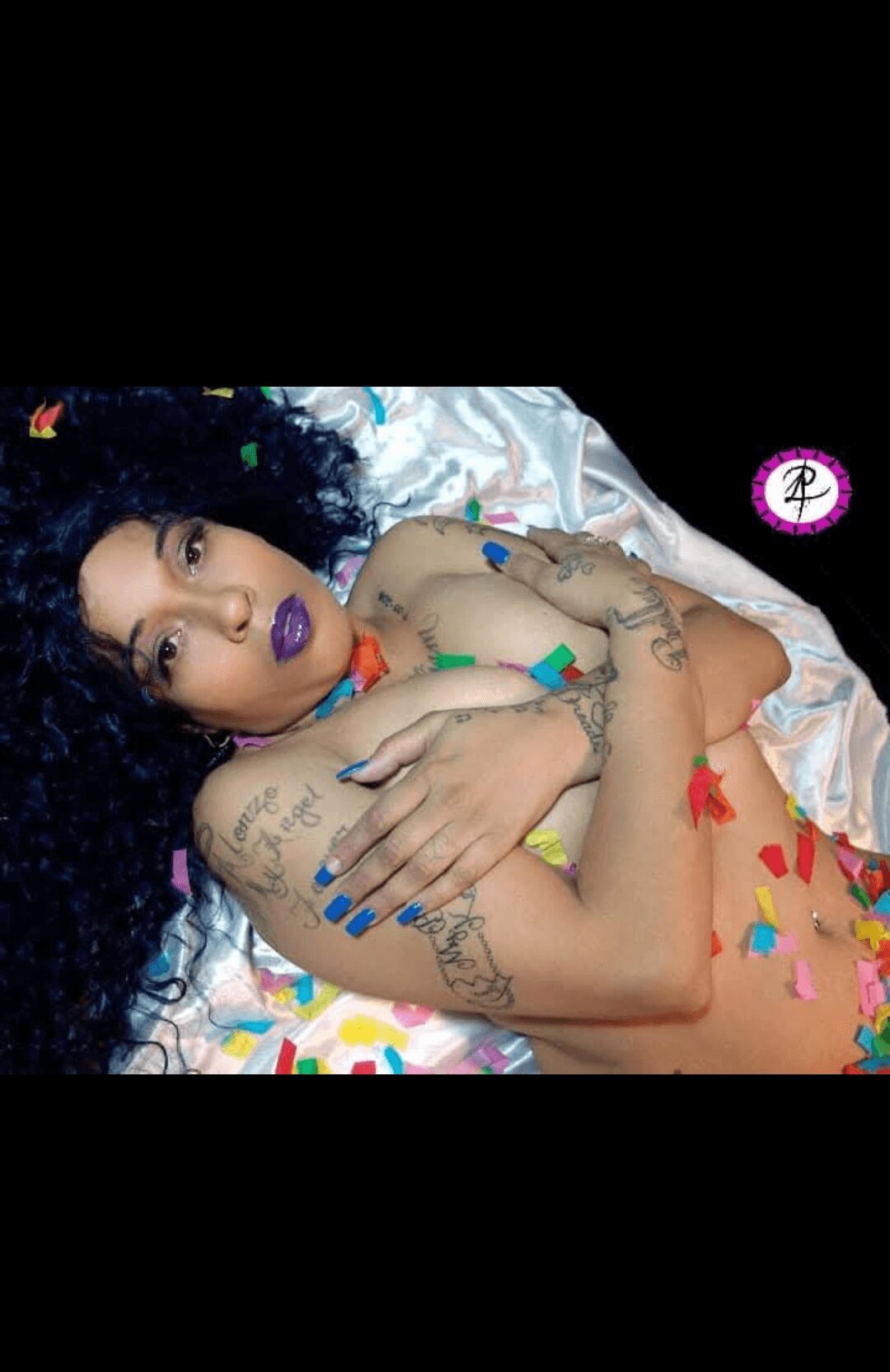 Person with curly hair lying on a bed with colorful flower petals, tattoos, and vibrant makeup, looking at the camera.