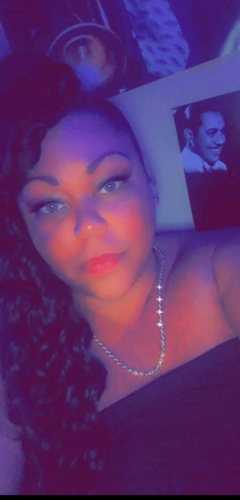 Woman with long curly hair wearing a necklace, lying down with a photo of a man in the background.