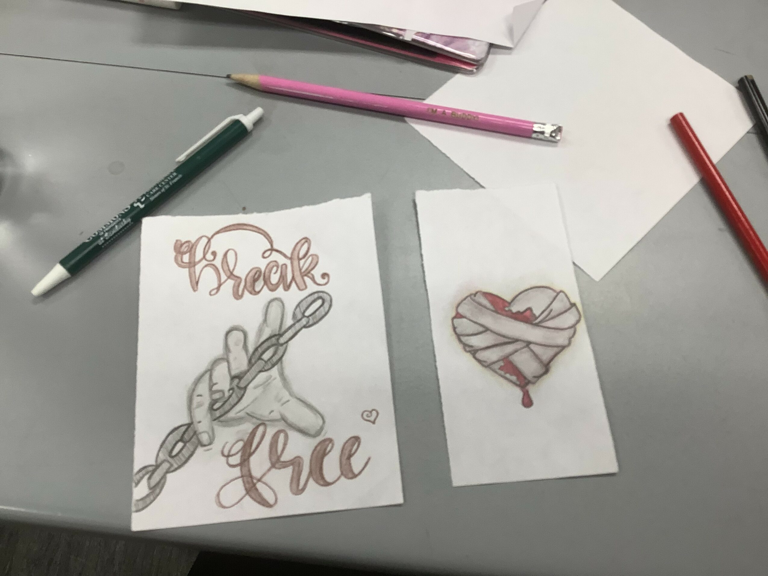 Two hand-drawn cards on a desk, one with a rope and the words 'break free', the other with a heart wrapped in bandages, surrounded by pens and papers.