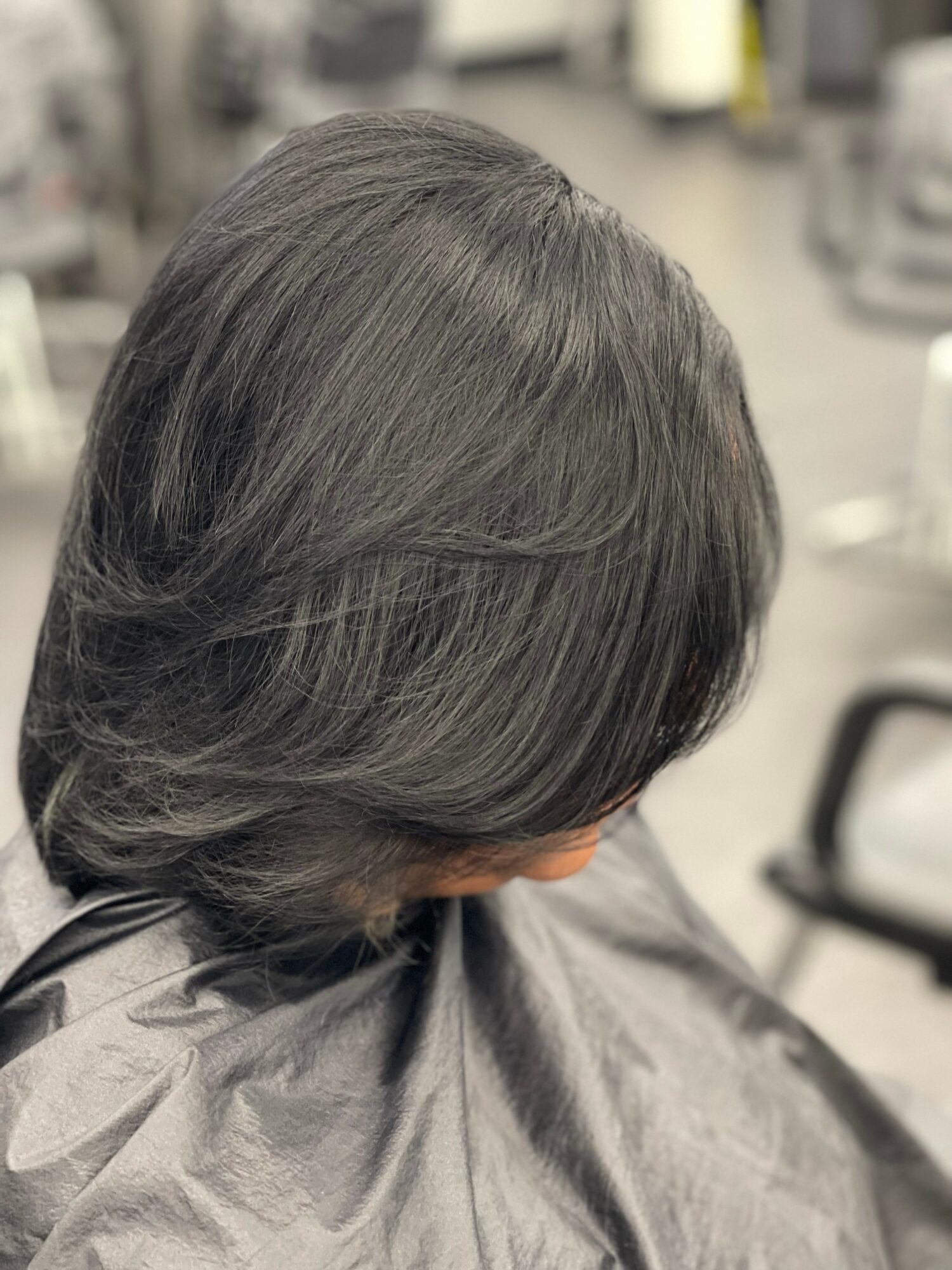 Side view of a person with shoulder-length dark hair, sitting in a salon chair, with blurred background.