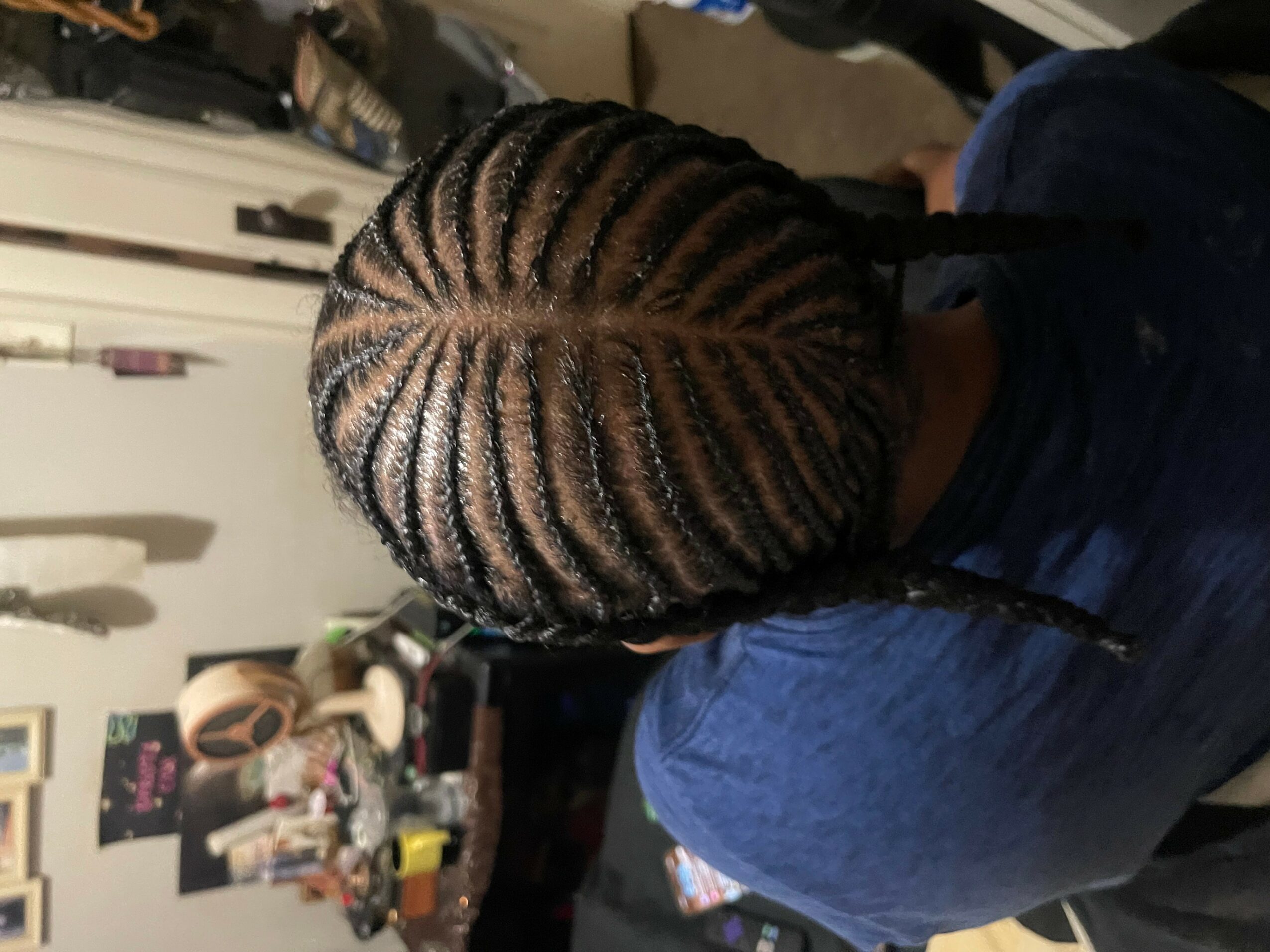 Top view of a person with braided hair, showing intricate cornrows, wearing a blue shirt, in a room with various items.