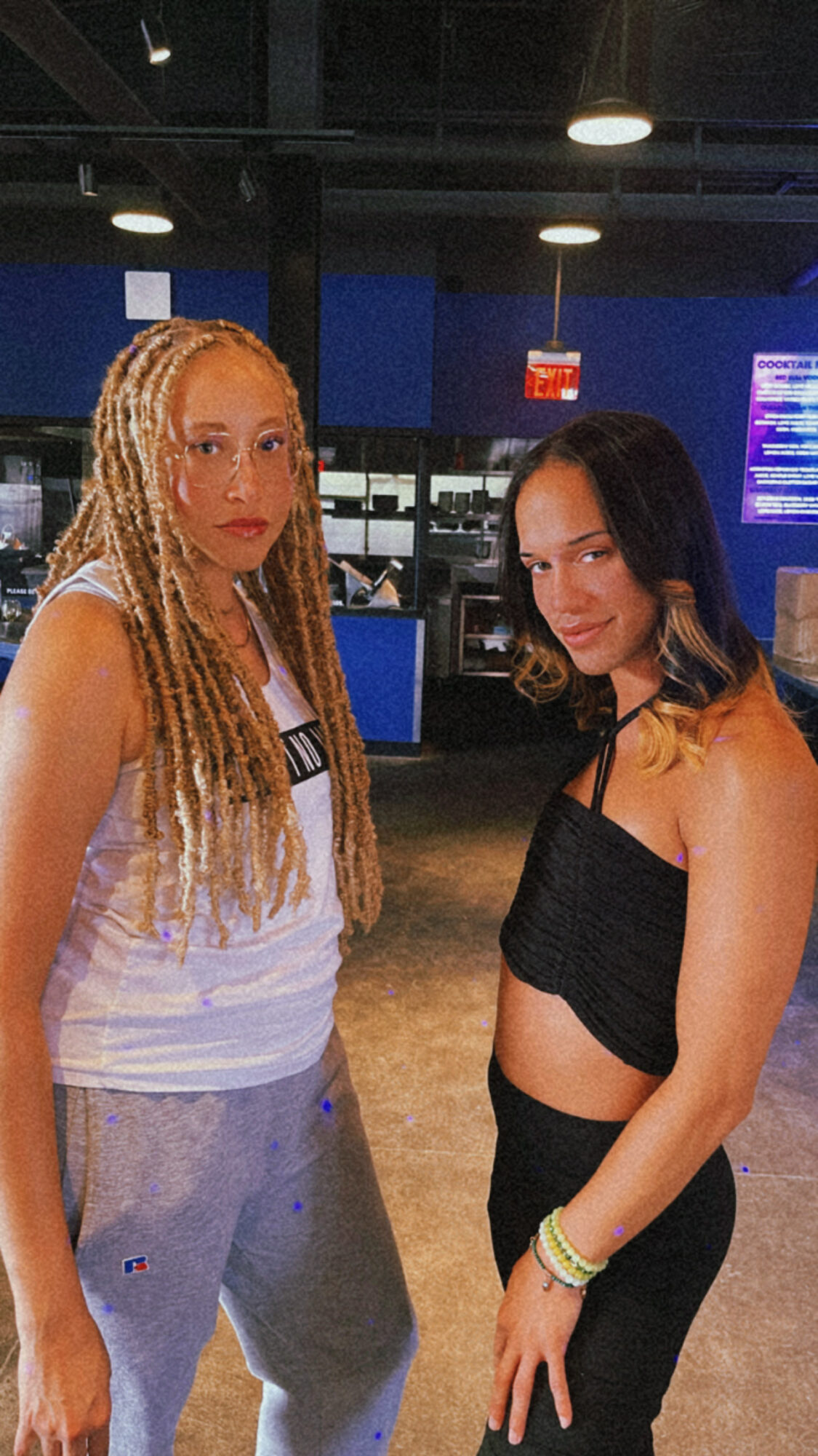 Two young women standing indoors, one with long blonde dreadlocks and the other with shoulder-length dark hair, both looking at the camera.